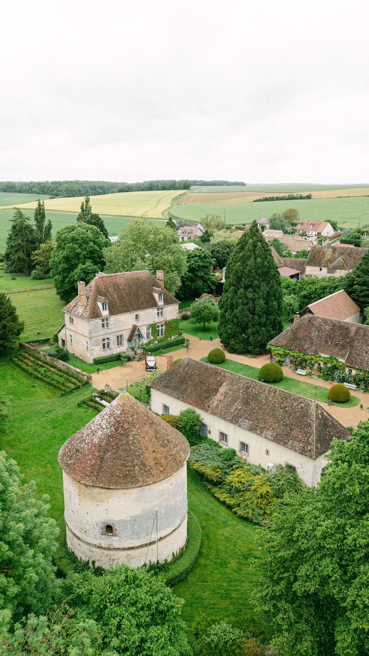 An aerial view of stone farm buildings and a round dovecote among green trees, with fields stretching into the distance.