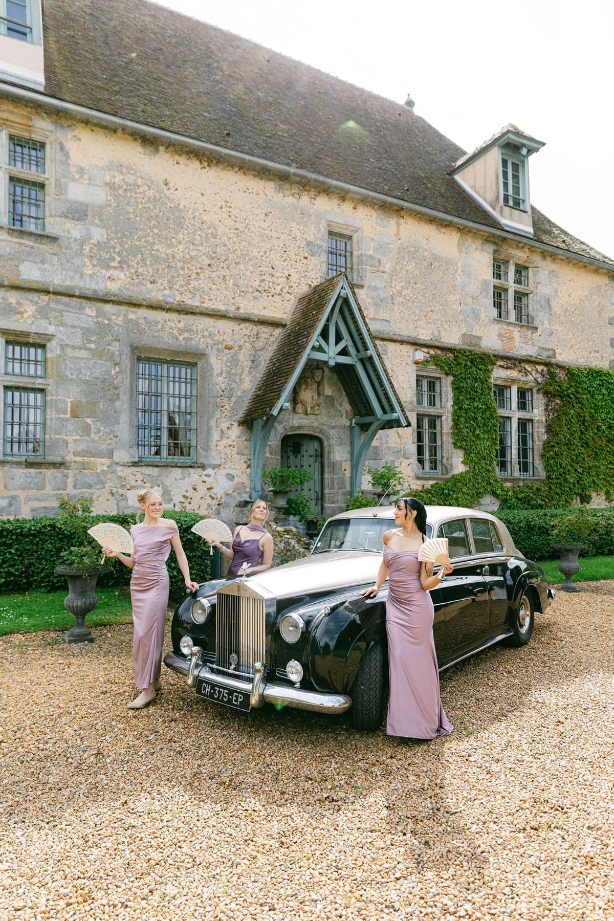 Three bridesmaids in mauve dresses stand around a black vintage car on gravel in front of a stone manor house.