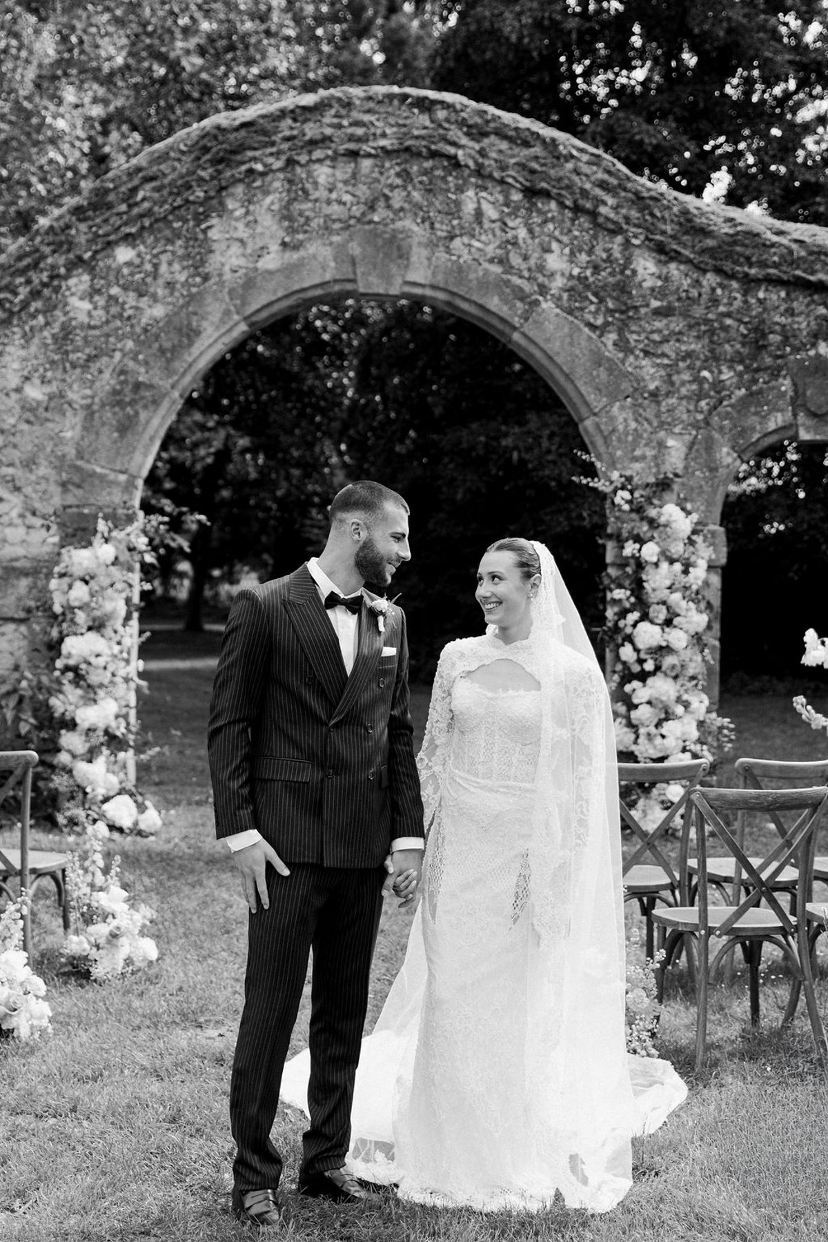 In black and white, the groom in a dark suit and the bride in a veil smile at each other near a stone arch.