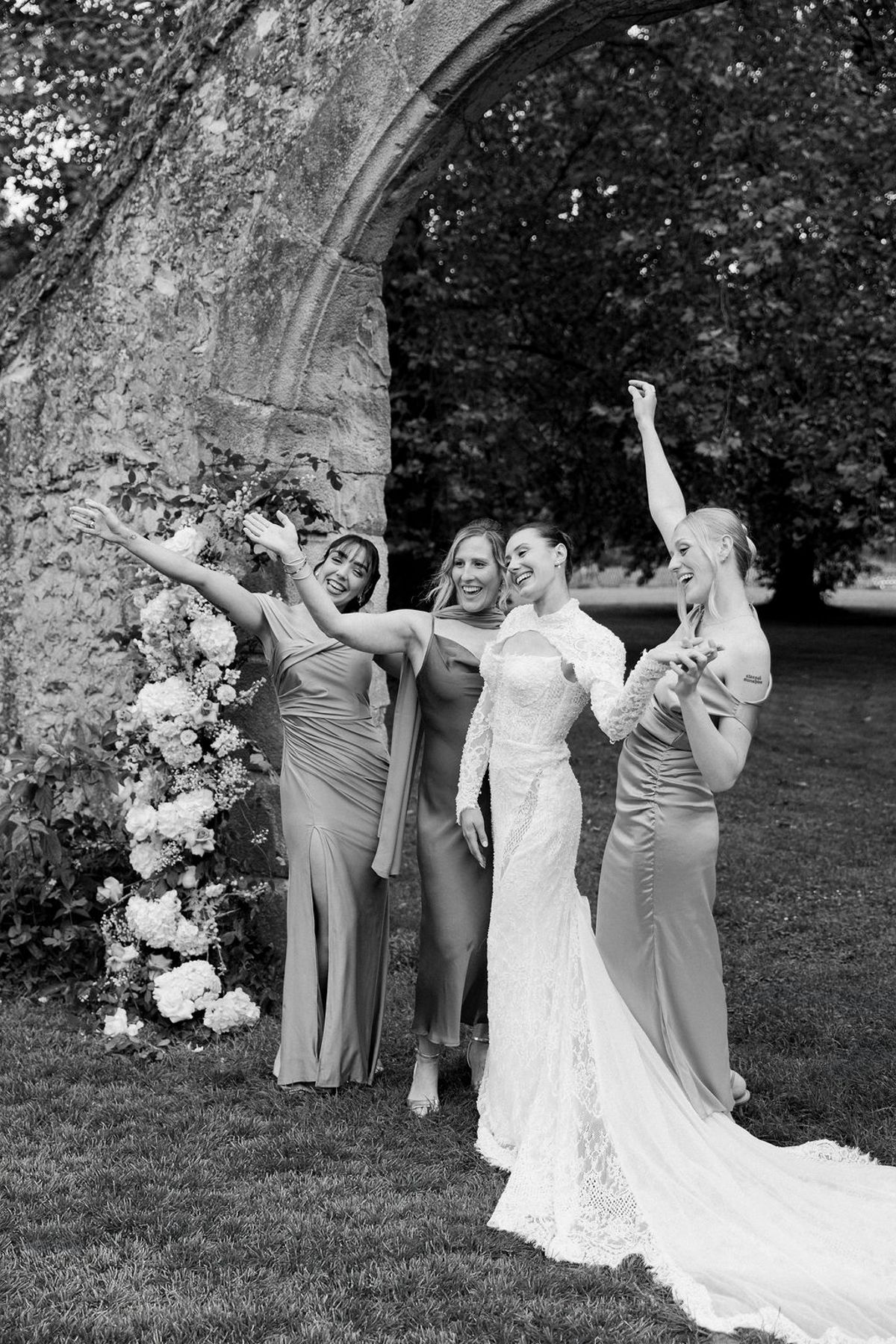 In black and white, the bride and her bridesmaids raise their arms and celebrate on a lawn in front of a ruined stone arch.