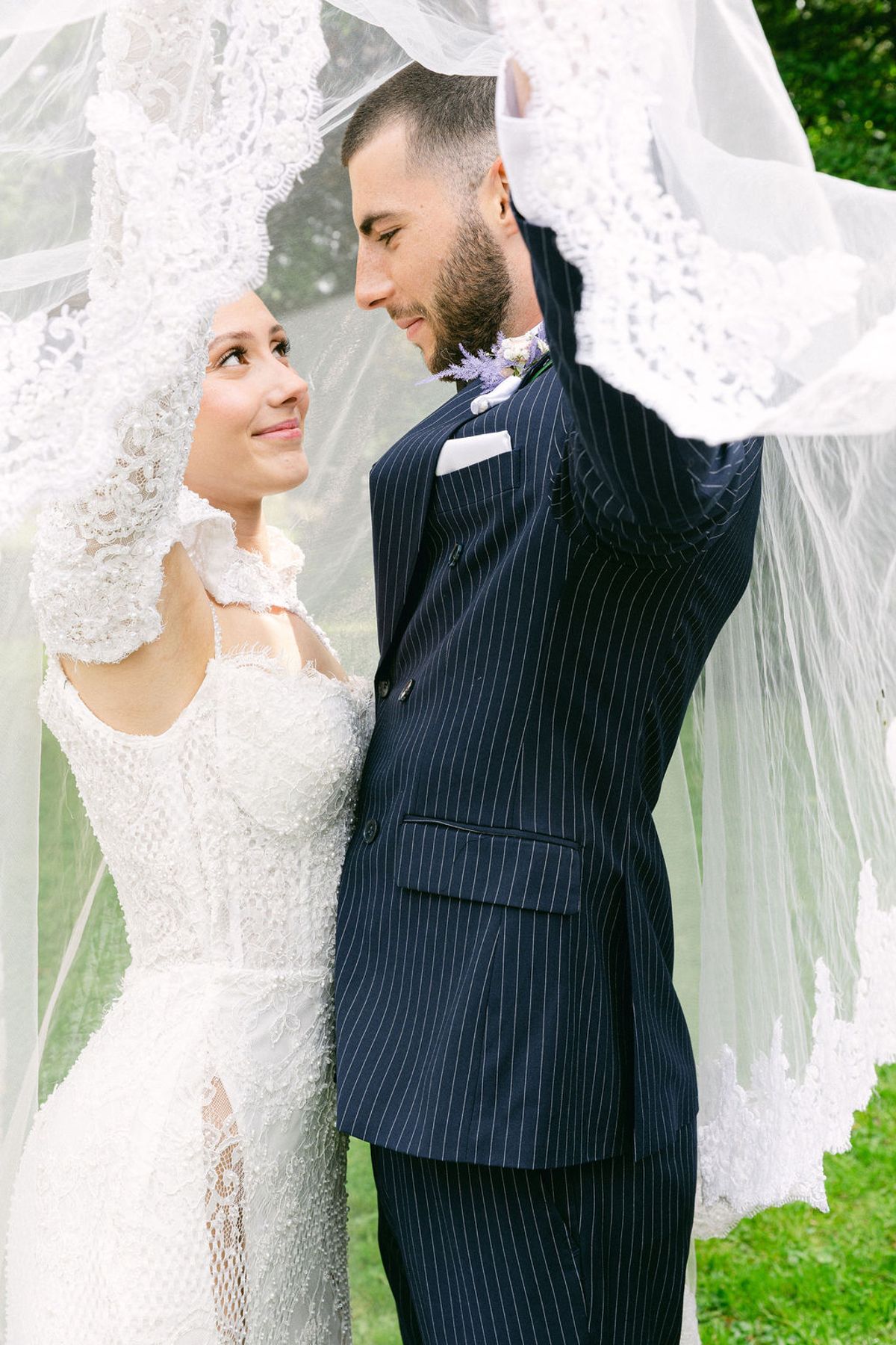The groom in a navy pinstriped suit lifts the bride's veil above them as the two smile at each other outdoors.