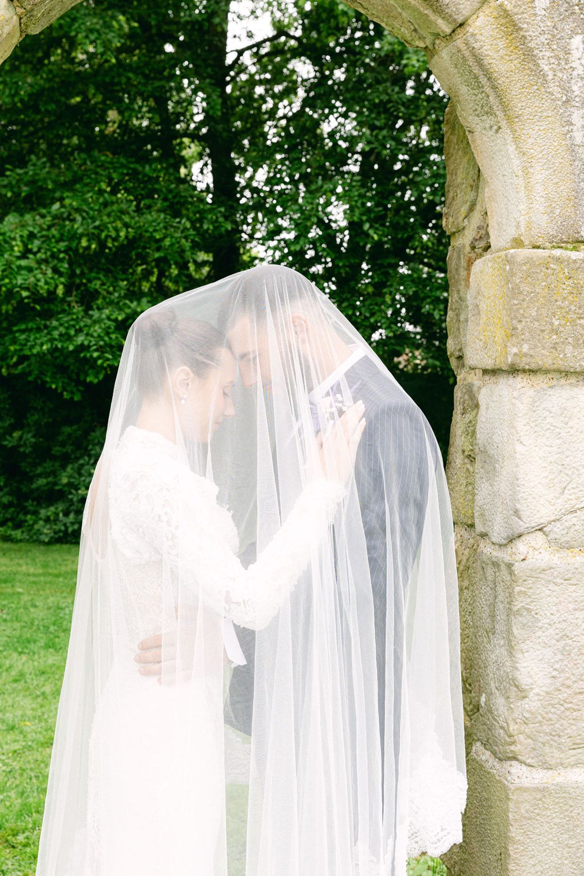 The couple stand close together beneath the bride's long white veil, beside a weathered stone archway with trees behind.