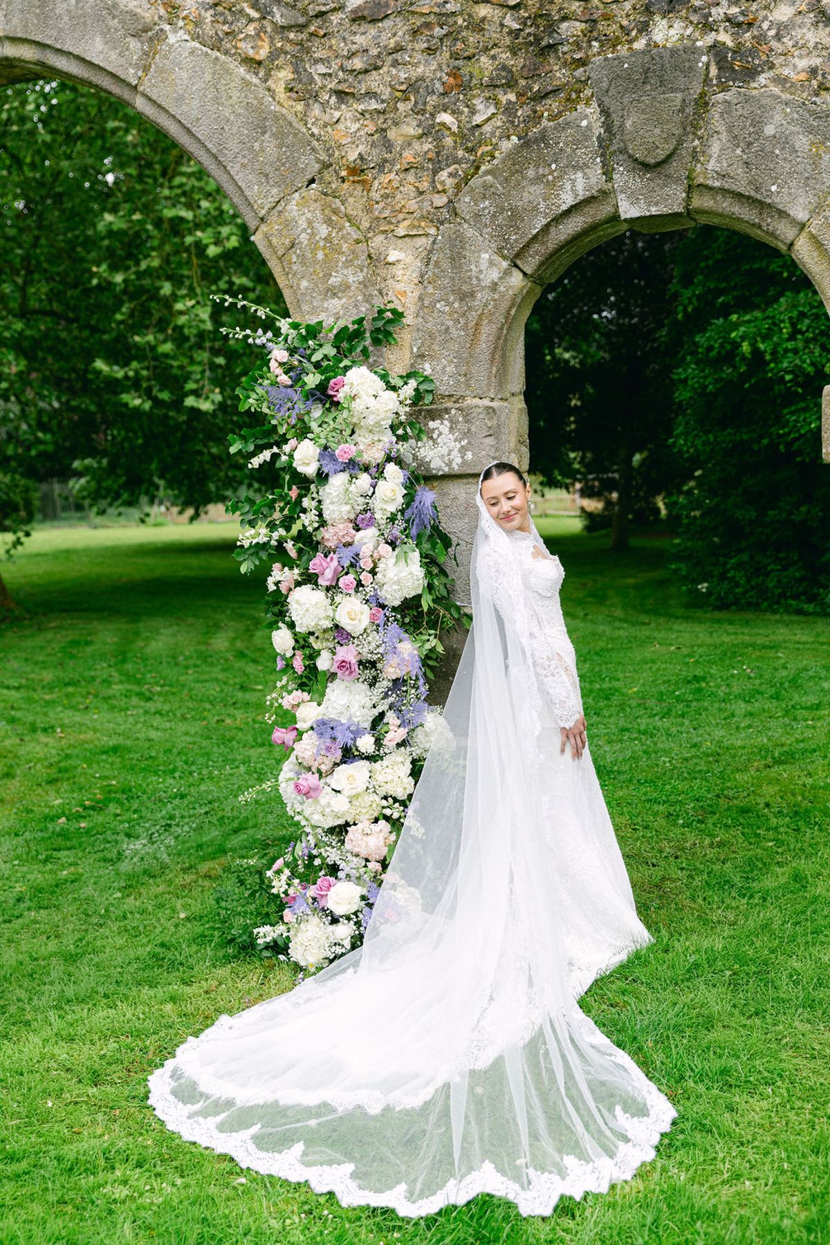 The bride in a long-sleeved lace gown and veil stands beside a tall flower arrangement under a stone arch on a lawn.