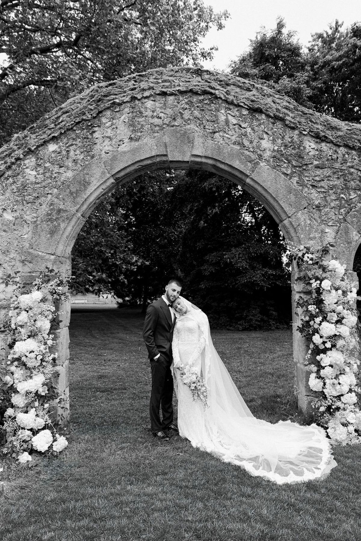 In black and white, the couple embrace beneath a stone arch decorated with flowers, the bride's long veil trailing on the grass.