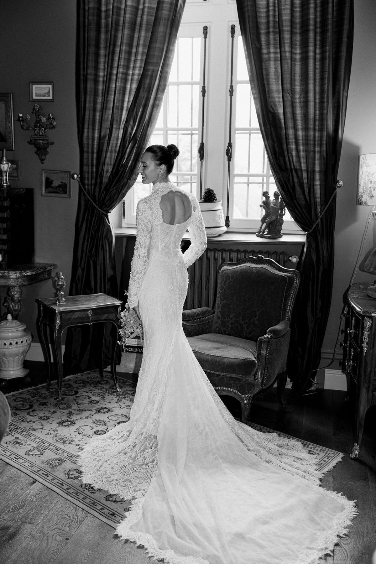 A black and white image of the bride seen from behind in a long lace gown, standing by a tall draped window in an ornate room.