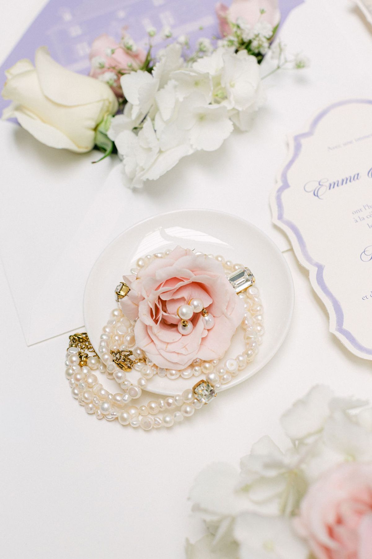 A pearl bracelet and pink rose on a small plate, laid beside a lilac-edged invitation and white flowers.