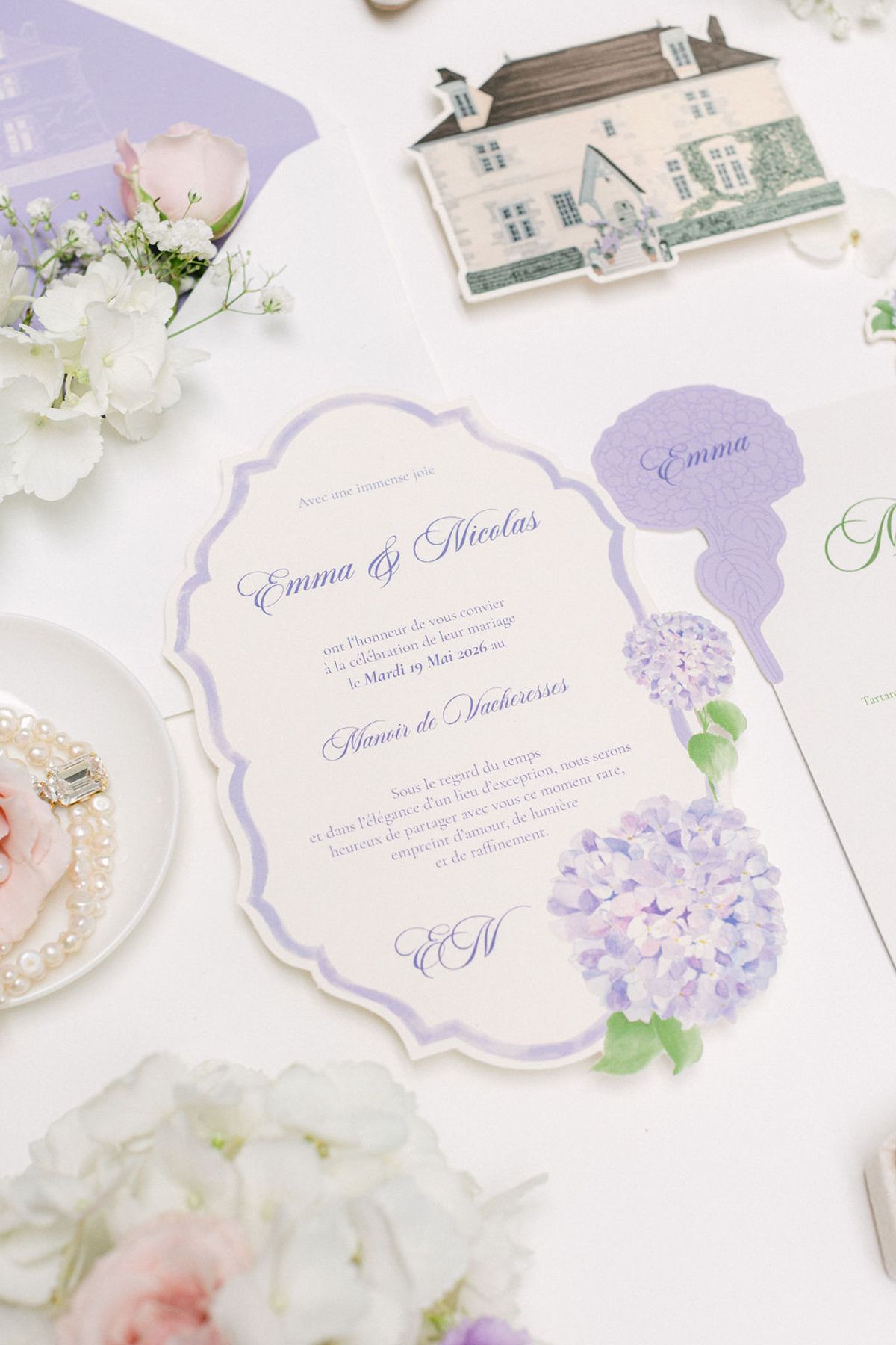 A flat lay of a scalloped lilac invitation card with a hydrangea illustration and a drawing of a chateau, beside white flowers.