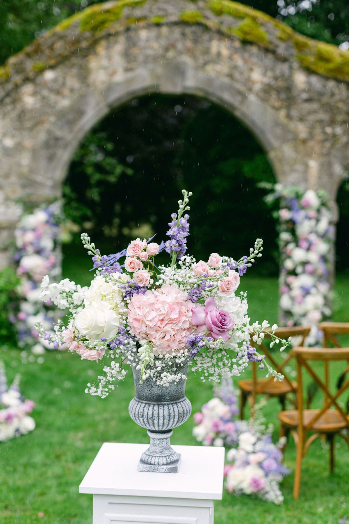 A large pink, white and lilac flower arrangement in a grey urn on a white plinth, with a mossy stone arch and wooden chairs behind.