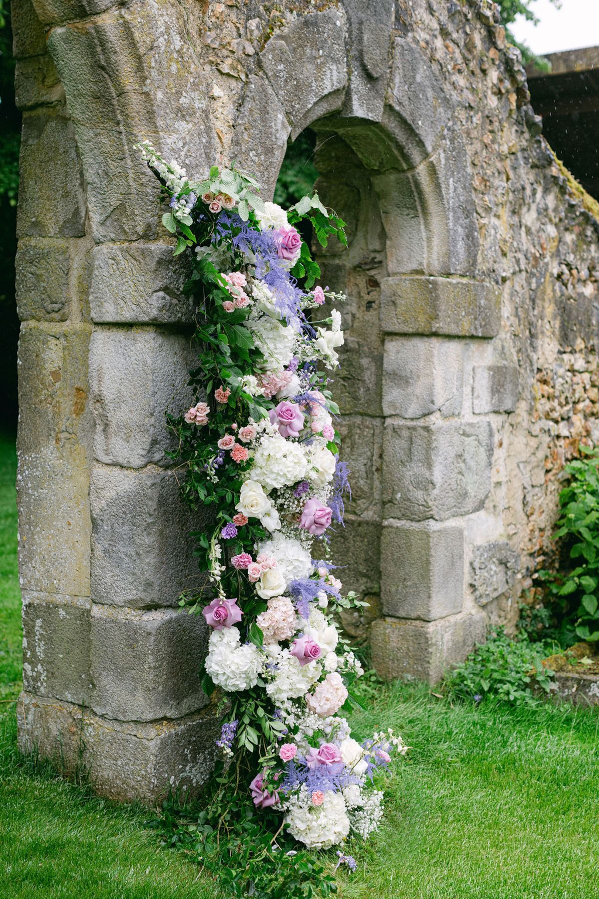 A tall column of white, pink and lilac flowers and greenery running up the side of an old stone archway.
