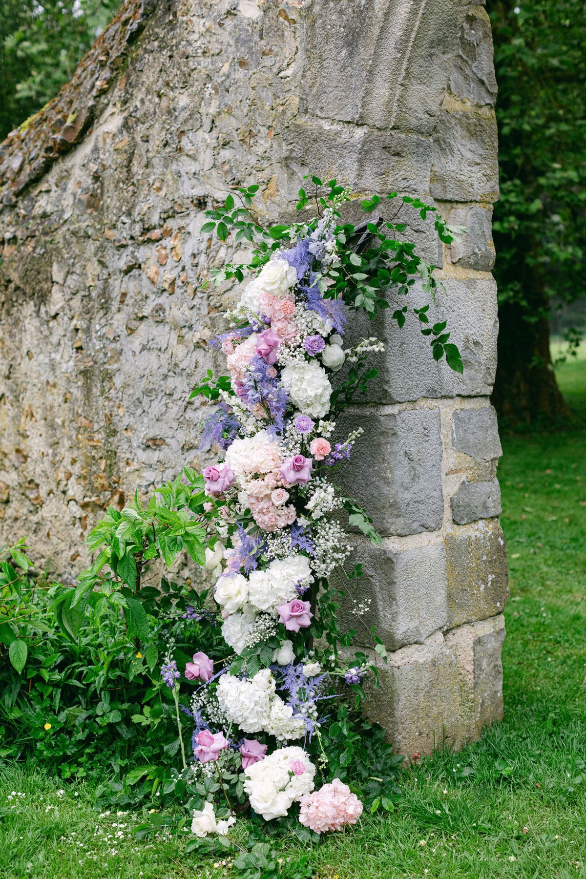 A tall arrangement of pink, white and lilac flowers rises against a weathered stone wall beside a green lawn.