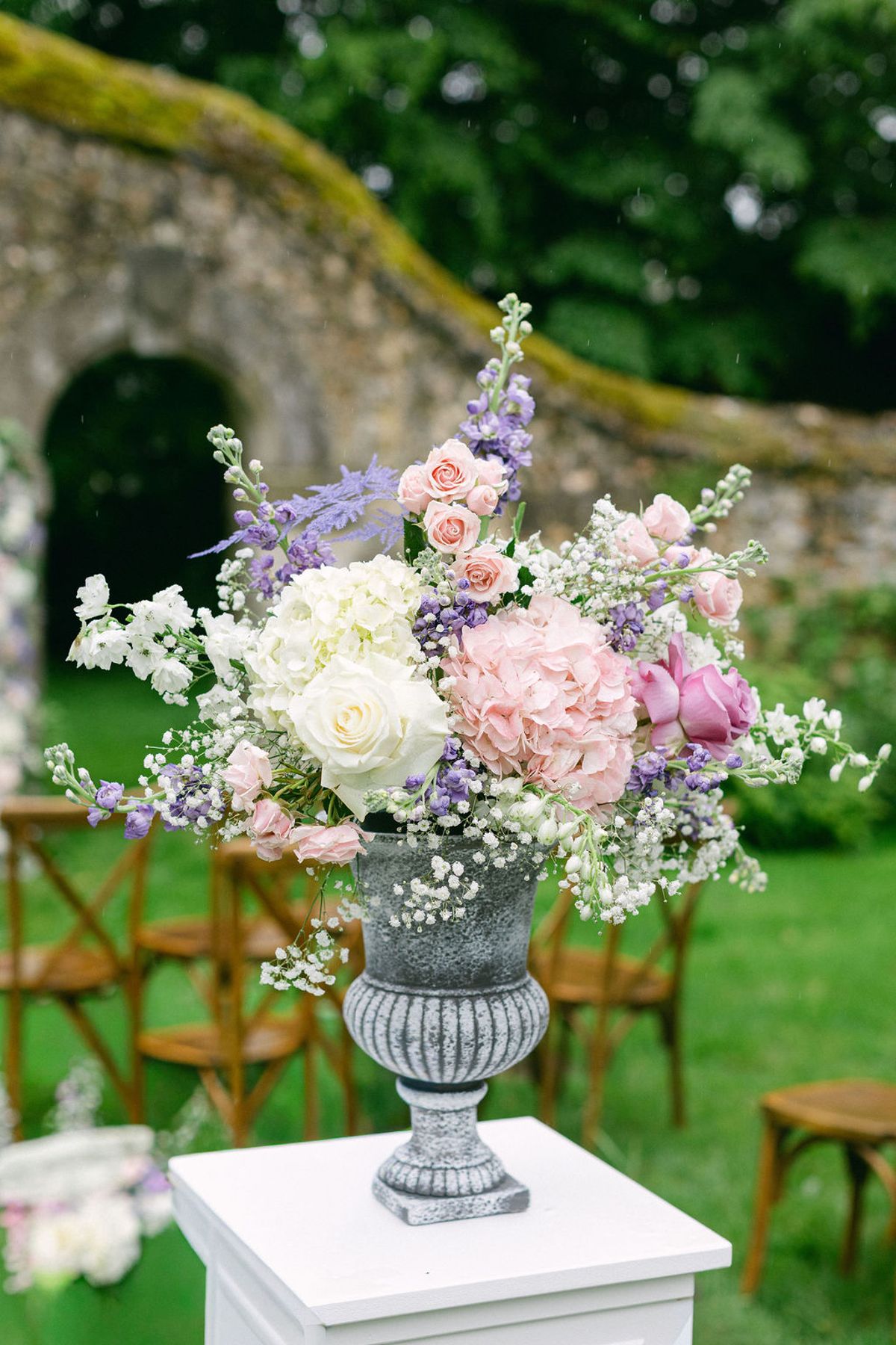 A large pink, white and lilac flower arrangement in a grey stone urn on a white plinth, with wooden chairs on the lawn behind.