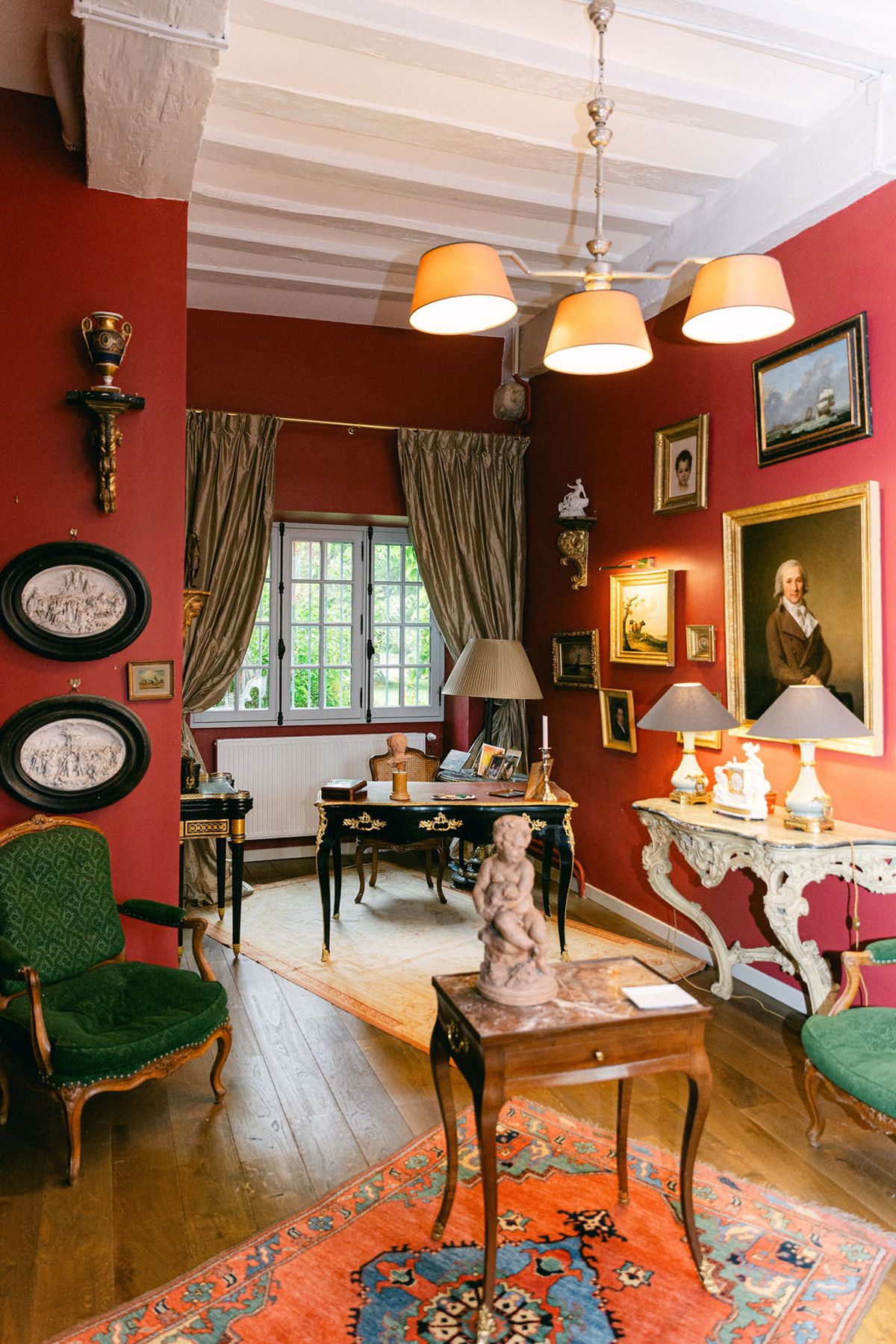 A room with red walls, framed portraits, a wooden desk, green armchairs, lit lamps and a colourful rug on a wooden floor.