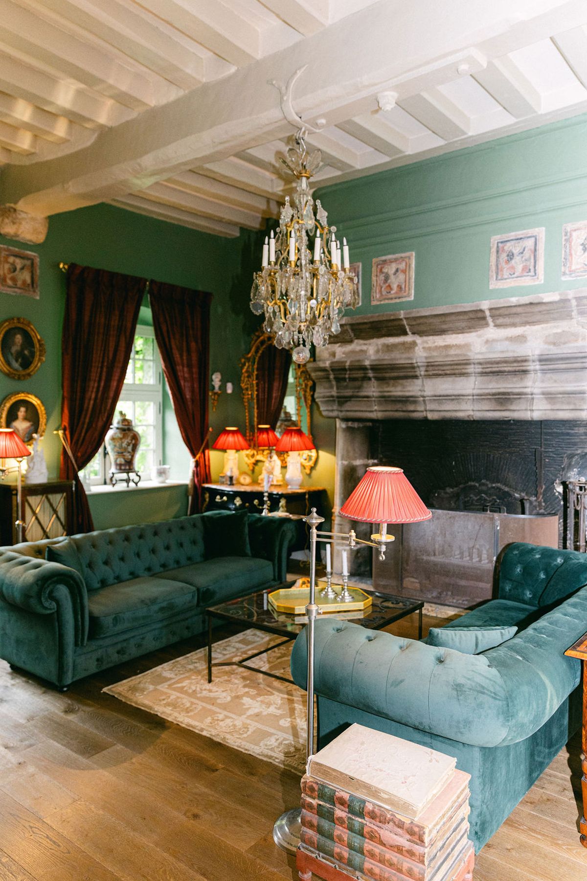 A sitting room with teal walls, green velvet sofas, a crystal chandelier, a large fireplace and red table lamps.