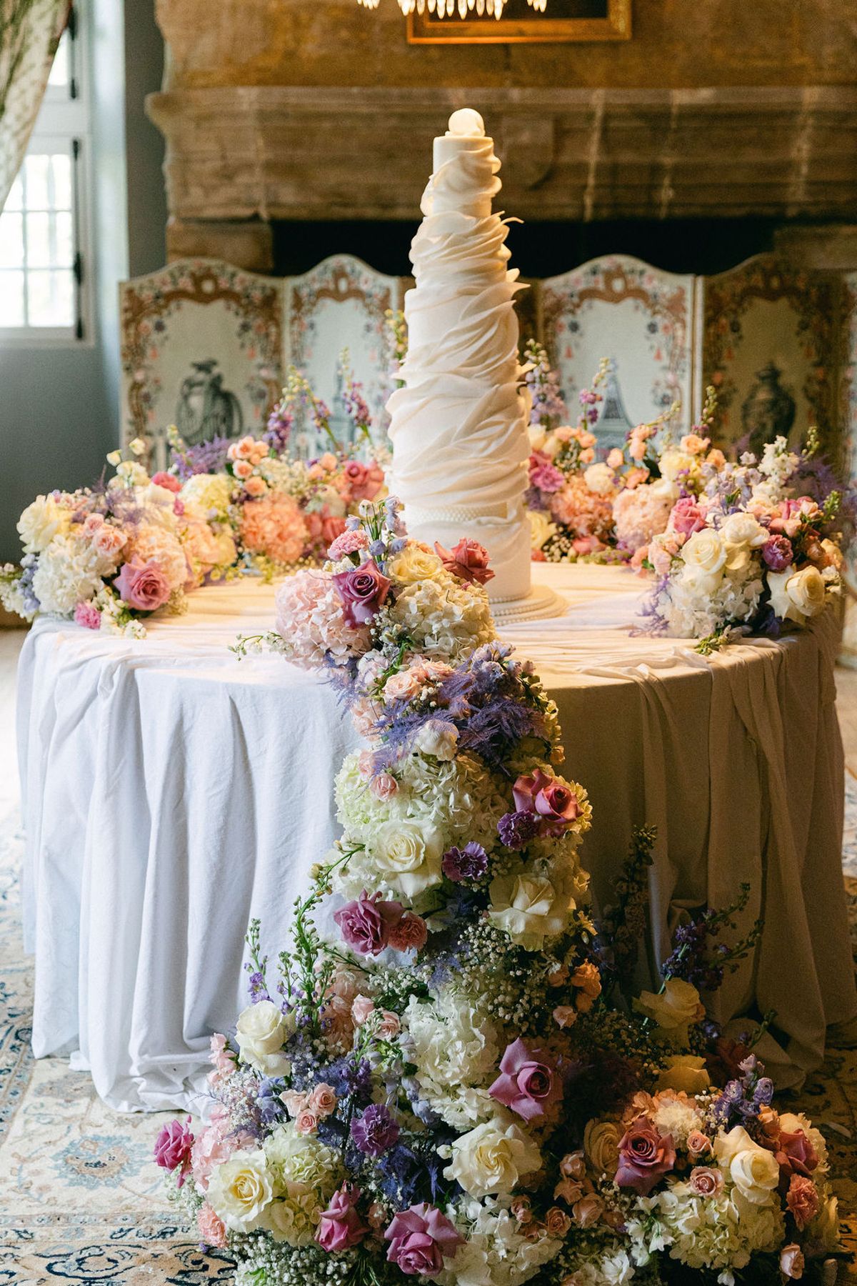 A tall, tiered white cake on a draped round table, surrounded by cascading pink, cream and lilac flowers in a stone-walled room.