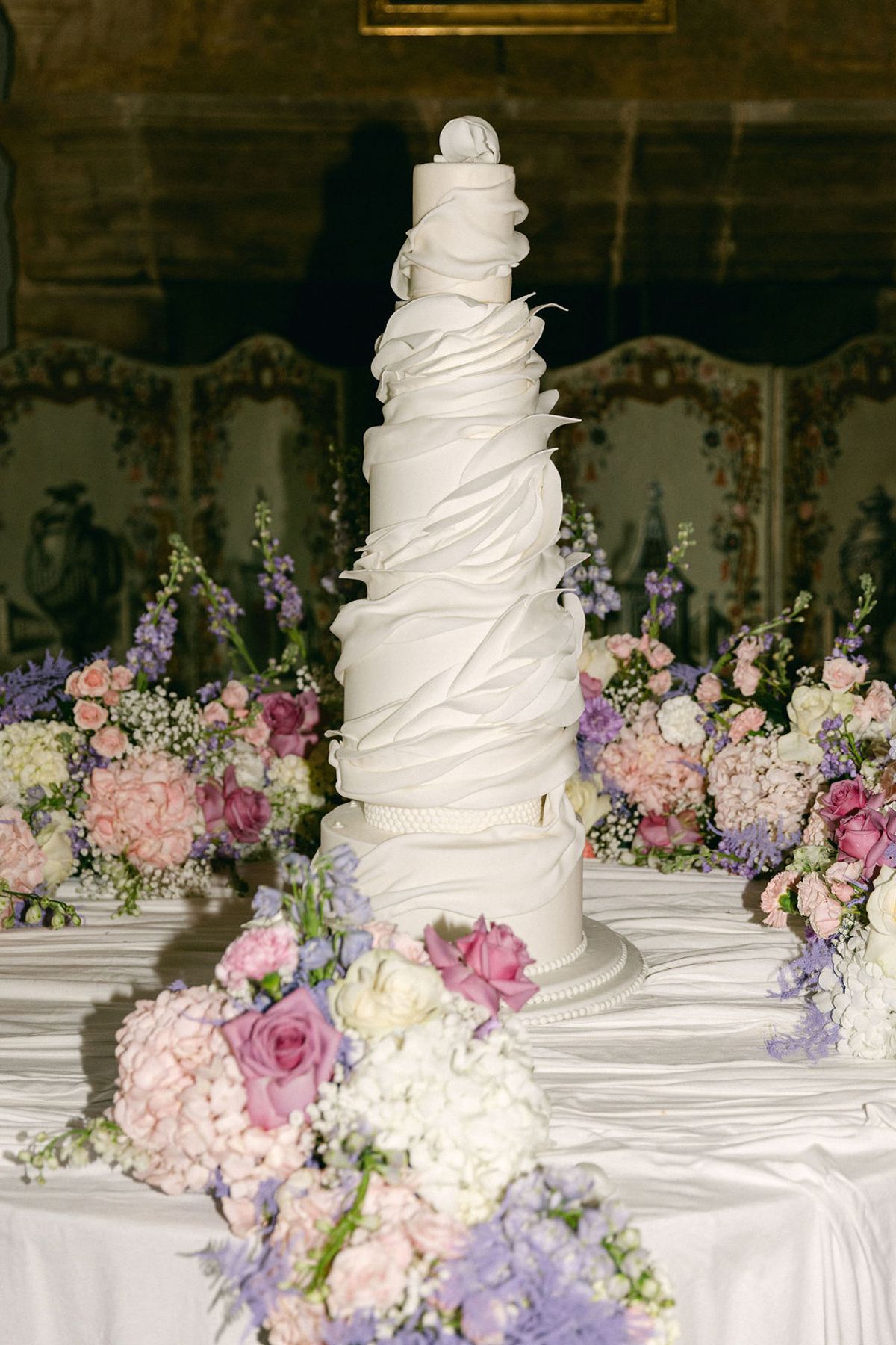 A tall white cake with draped, ruffled tiers stands on a table surrounded by pink, lilac and white flowers in a decorated room.
