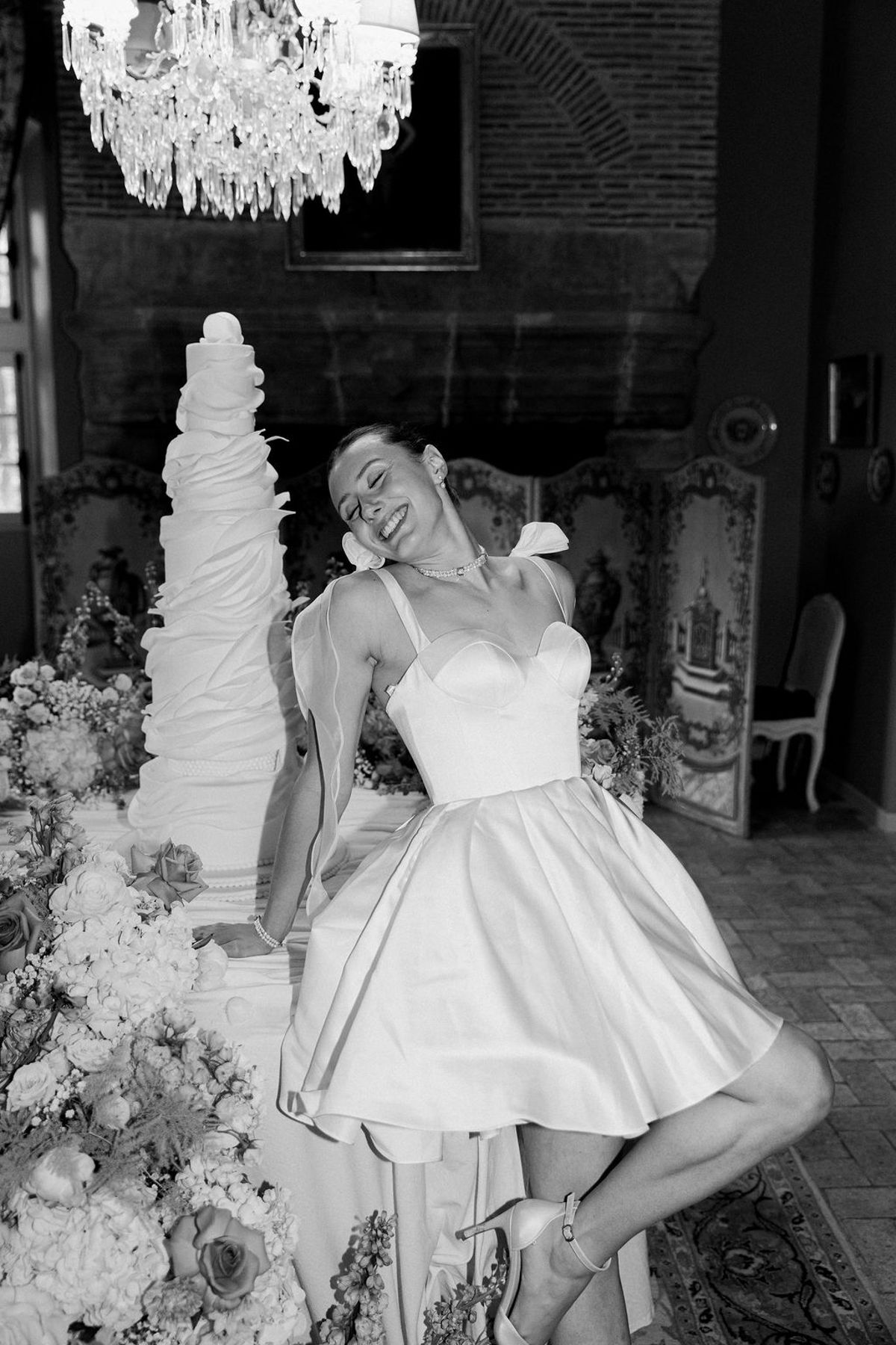 In black and white, the bride in a short satin dress leans playfully against a table beside a very tall cake, under a chandelier.