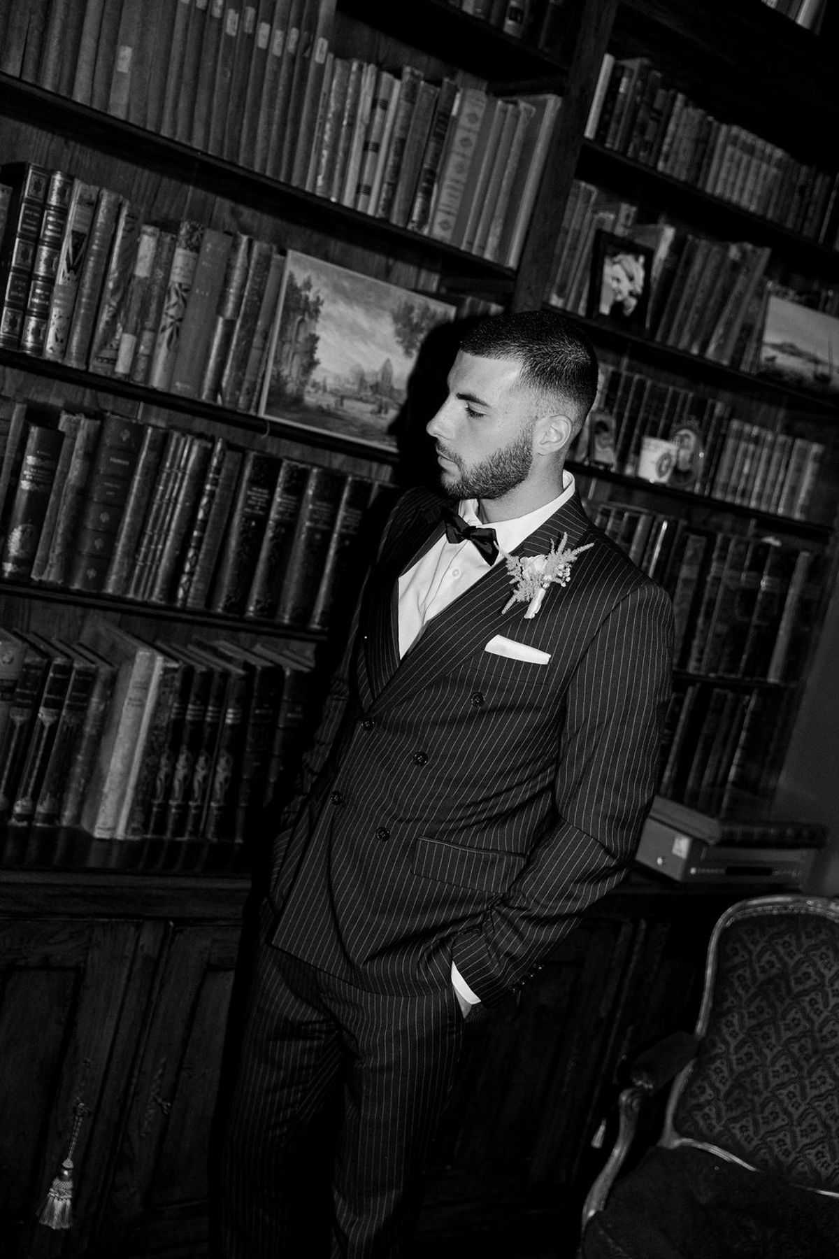 In black and white, the groom in a pinstriped suit stands with his hands in his pockets before shelves of old books.