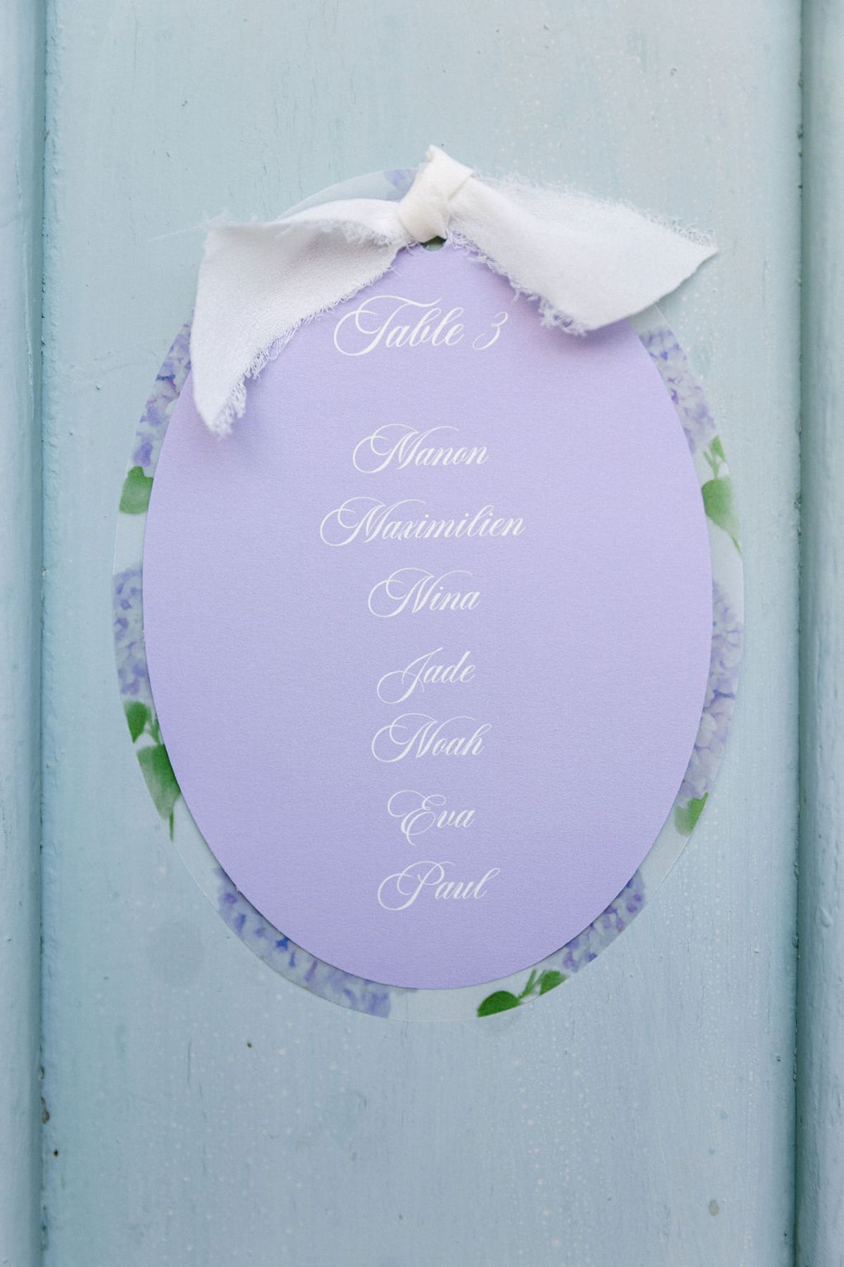 A lilac oval sign with a white ribbon bow and handwritten script lists names, and is fixed to a pale blue door.