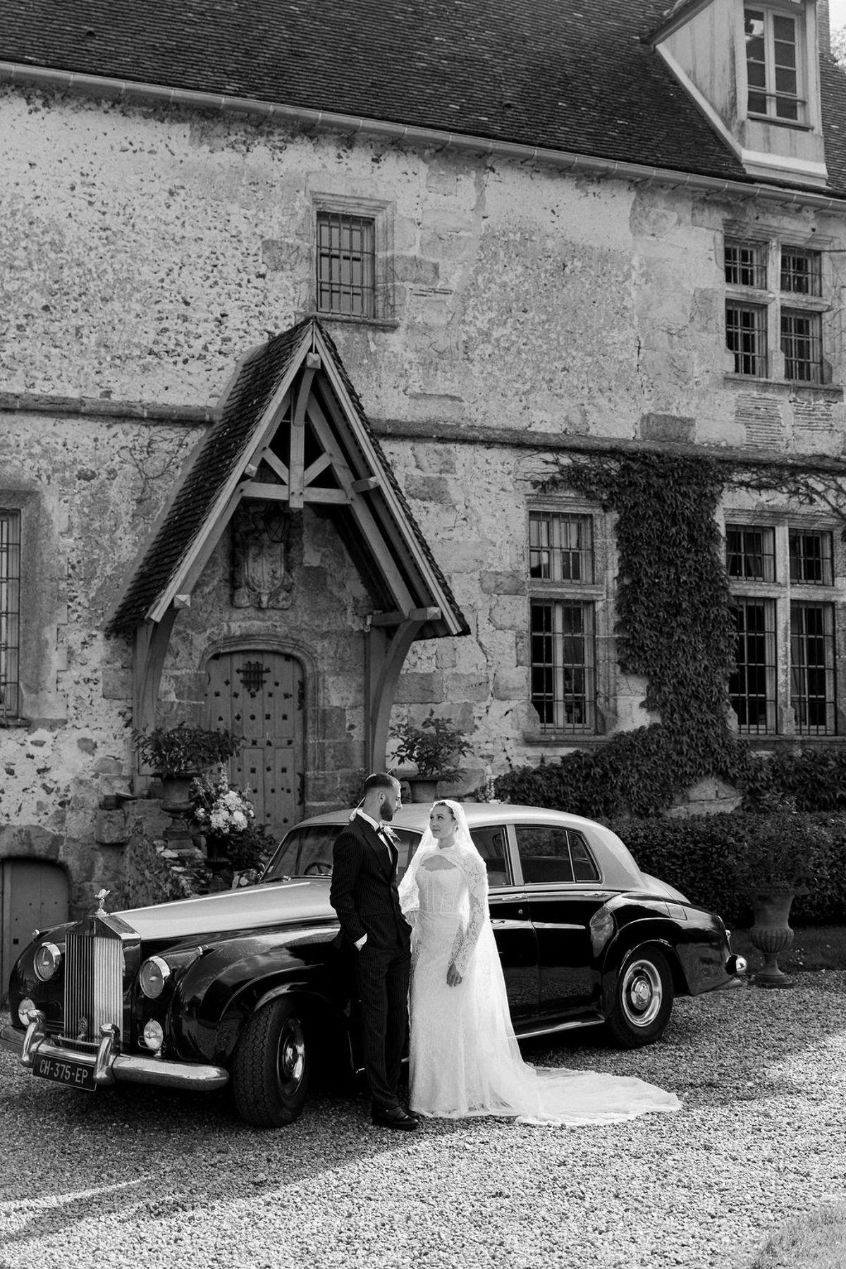 In black and white, the couple stand beside a vintage car on gravel in front of a stone chateau with a timber porch.