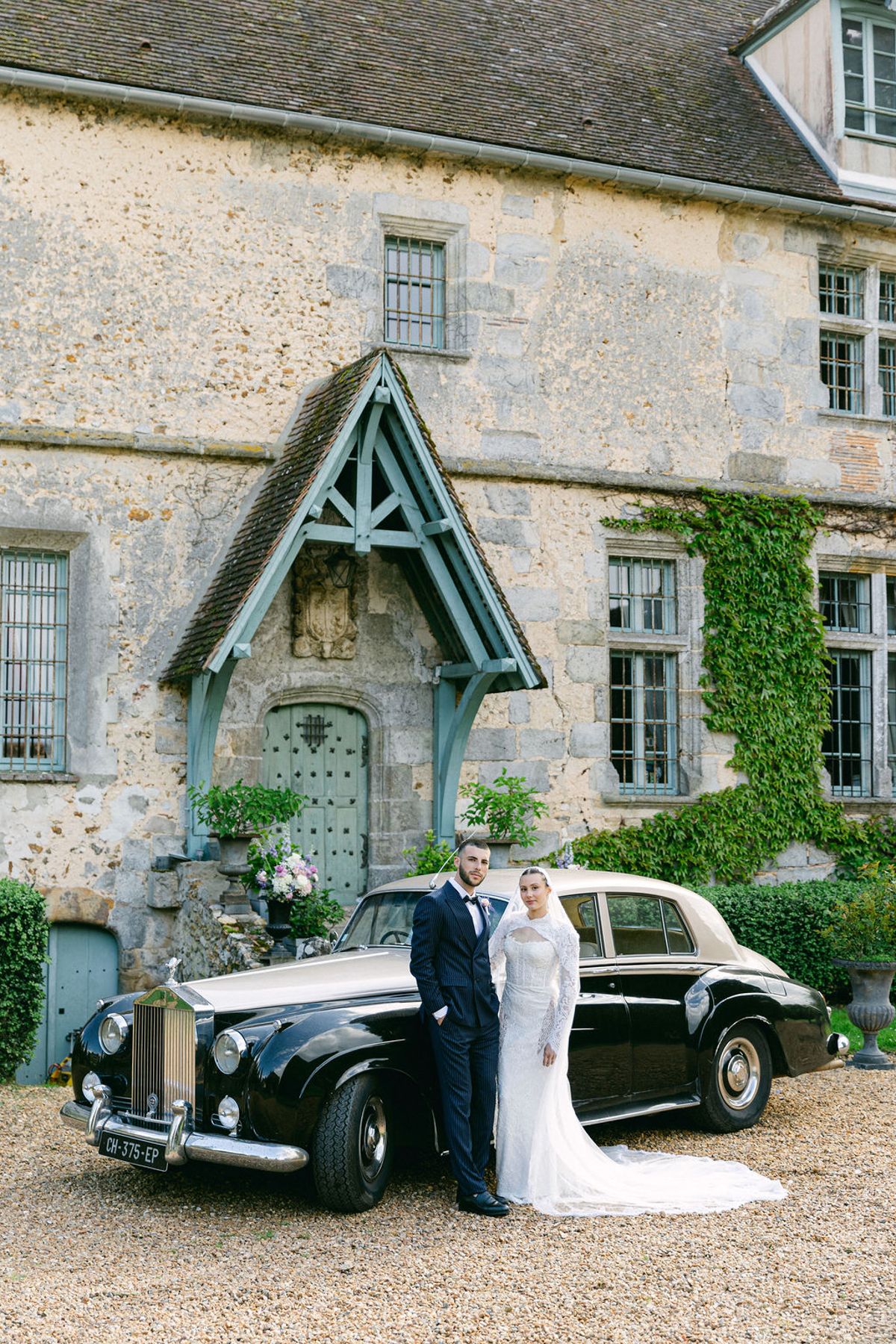 The couple stand together beside a black vintage car on gravel, small against a large stone manor house with ivy.