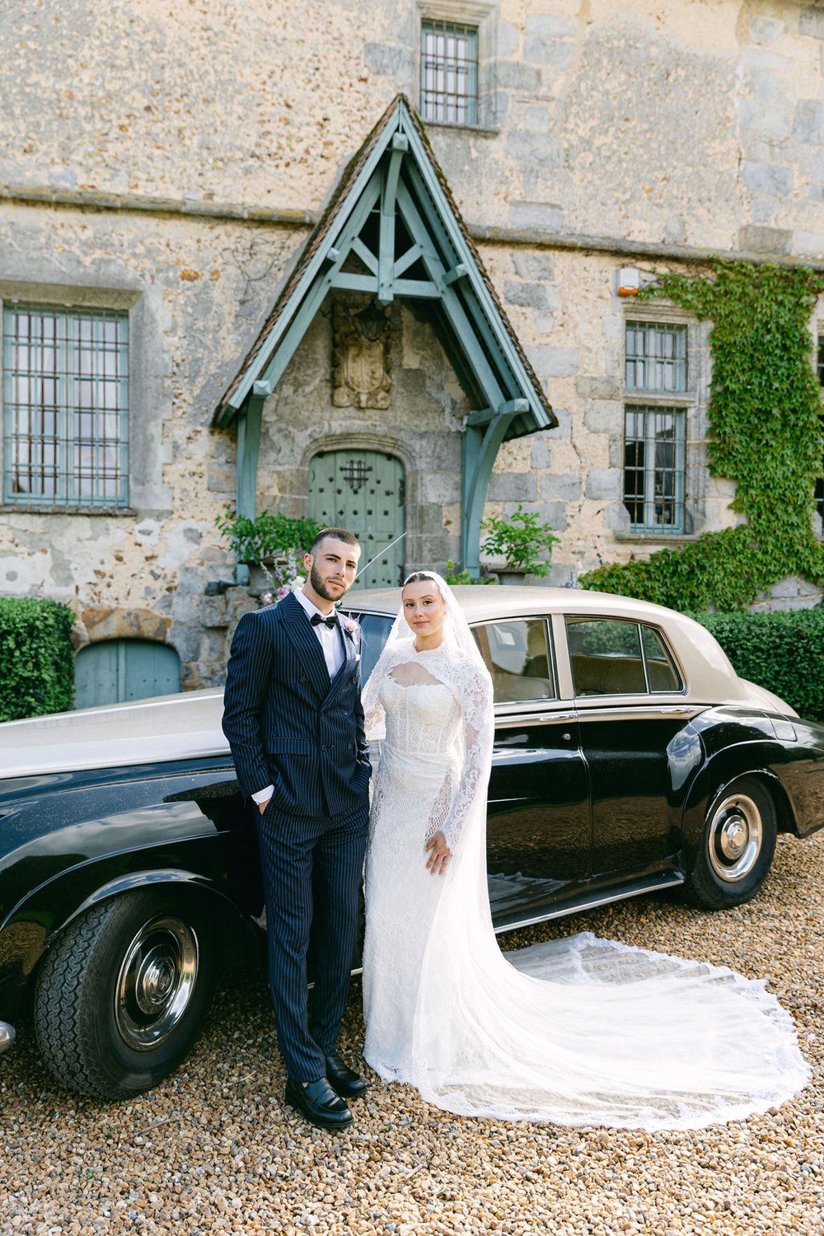 The couple stand arm in arm beside a black vintage car on gravel, in front of a stone building with a pale blue porch.
