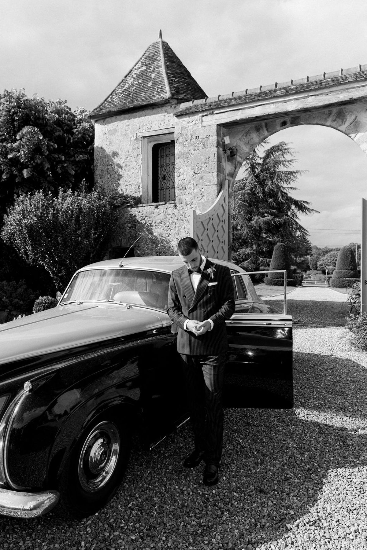 A black and white image of the groom standing with his head bowed beside a vintage car, with a stone tower and archway behind.