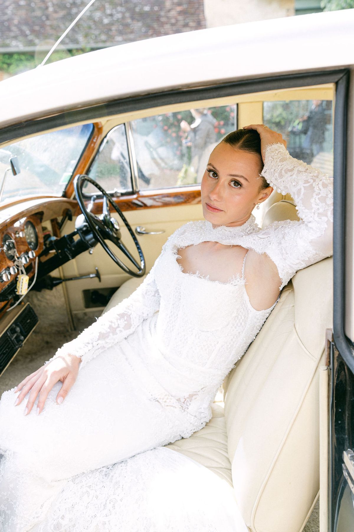 The bride in a white lace gown reclines on the cream leather seat of a vintage car, looking towards the camera.