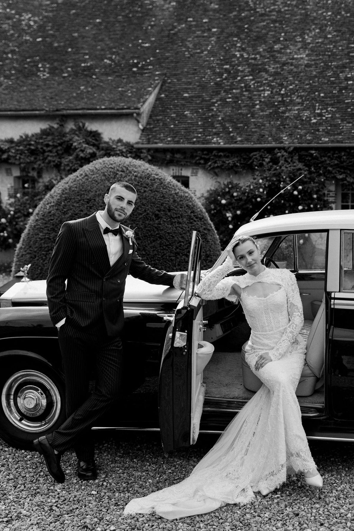 In black and white, the groom leans against a vintage car while the bride sits in its open door beside him.