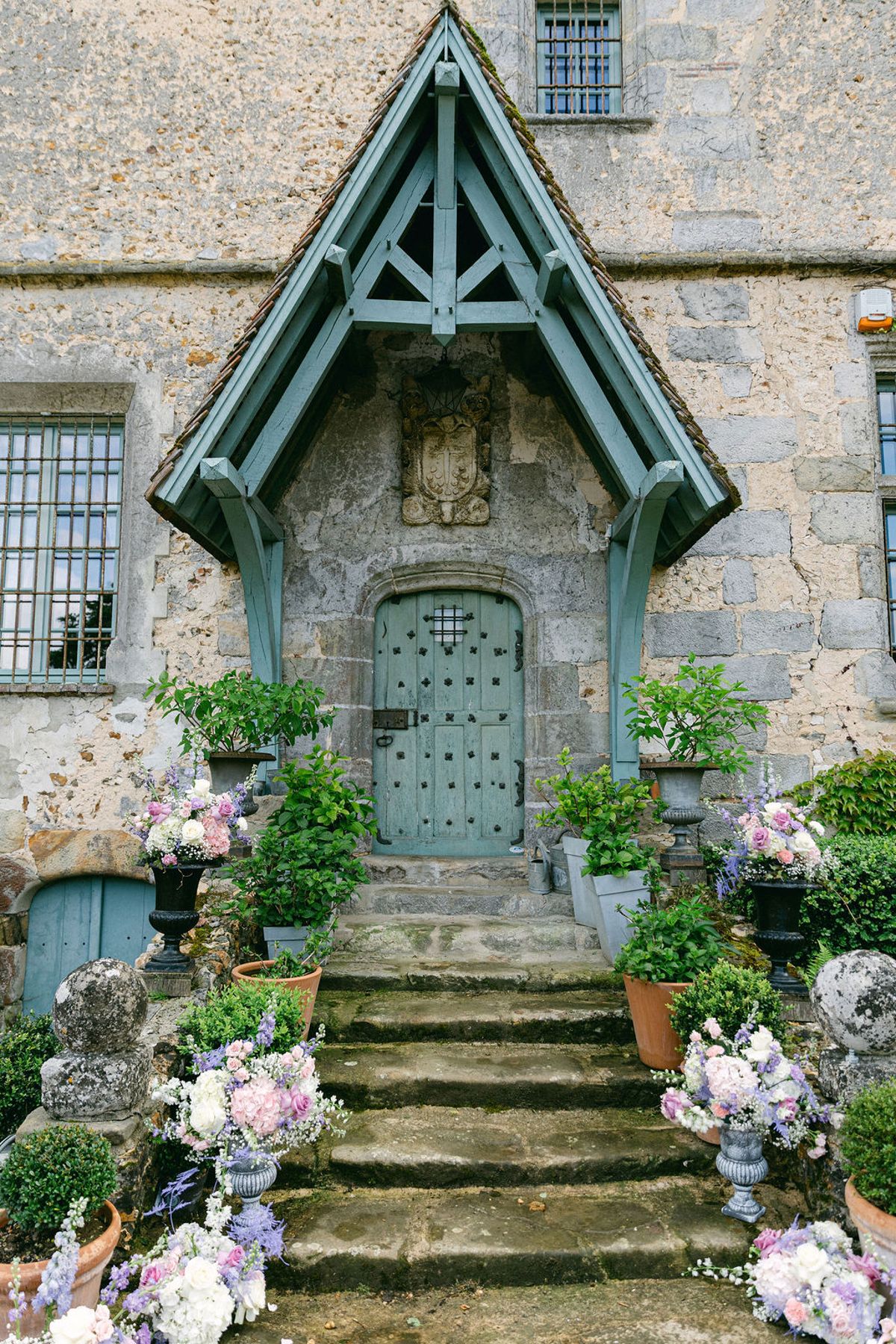 A stone chateau entrance with a pale blue door and timber porch, its stone steps lined with pots of pastel flowers.