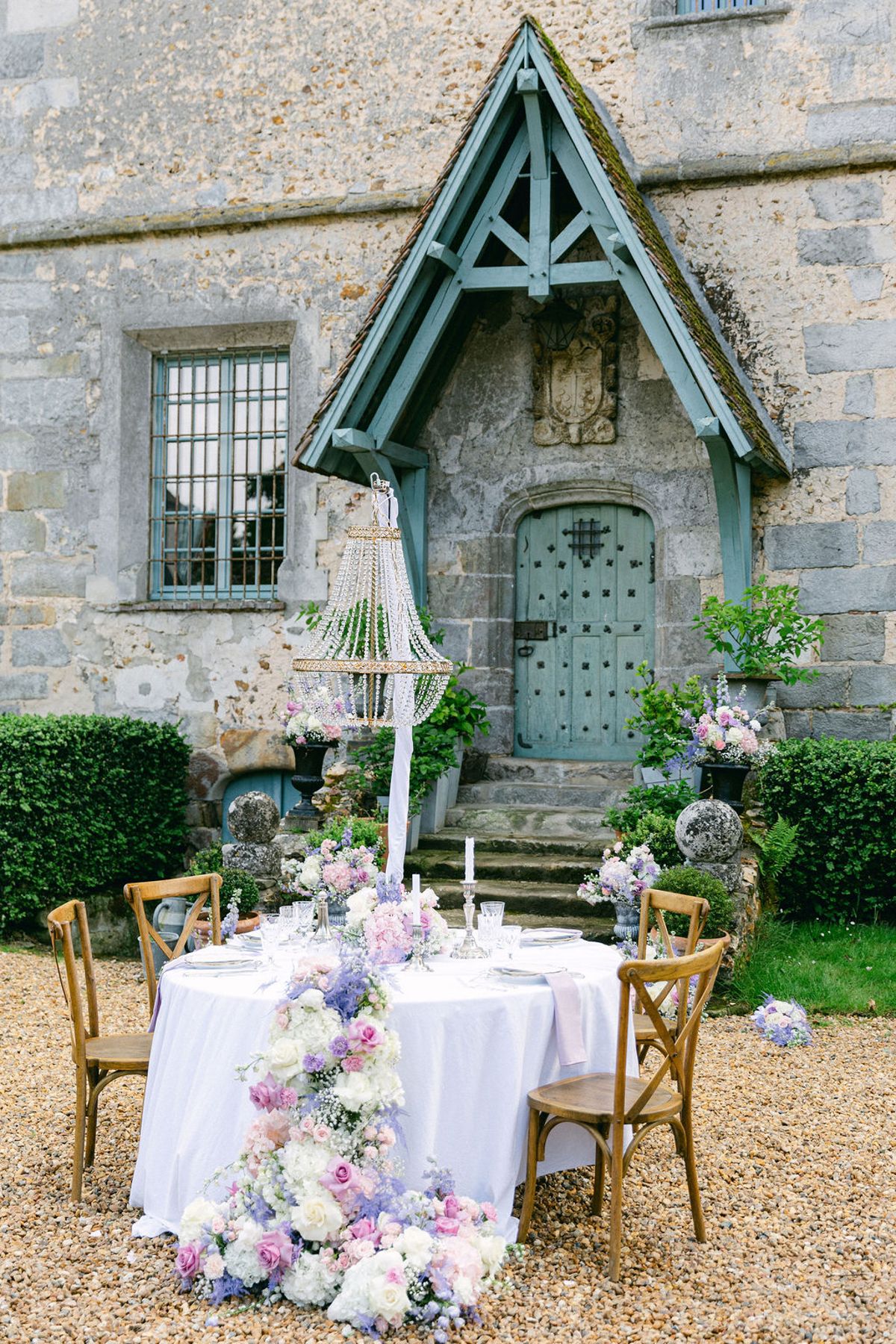 A round table with white cloth, wooden chairs and a cascade of pastel flowers set on gravel before a stone porch.