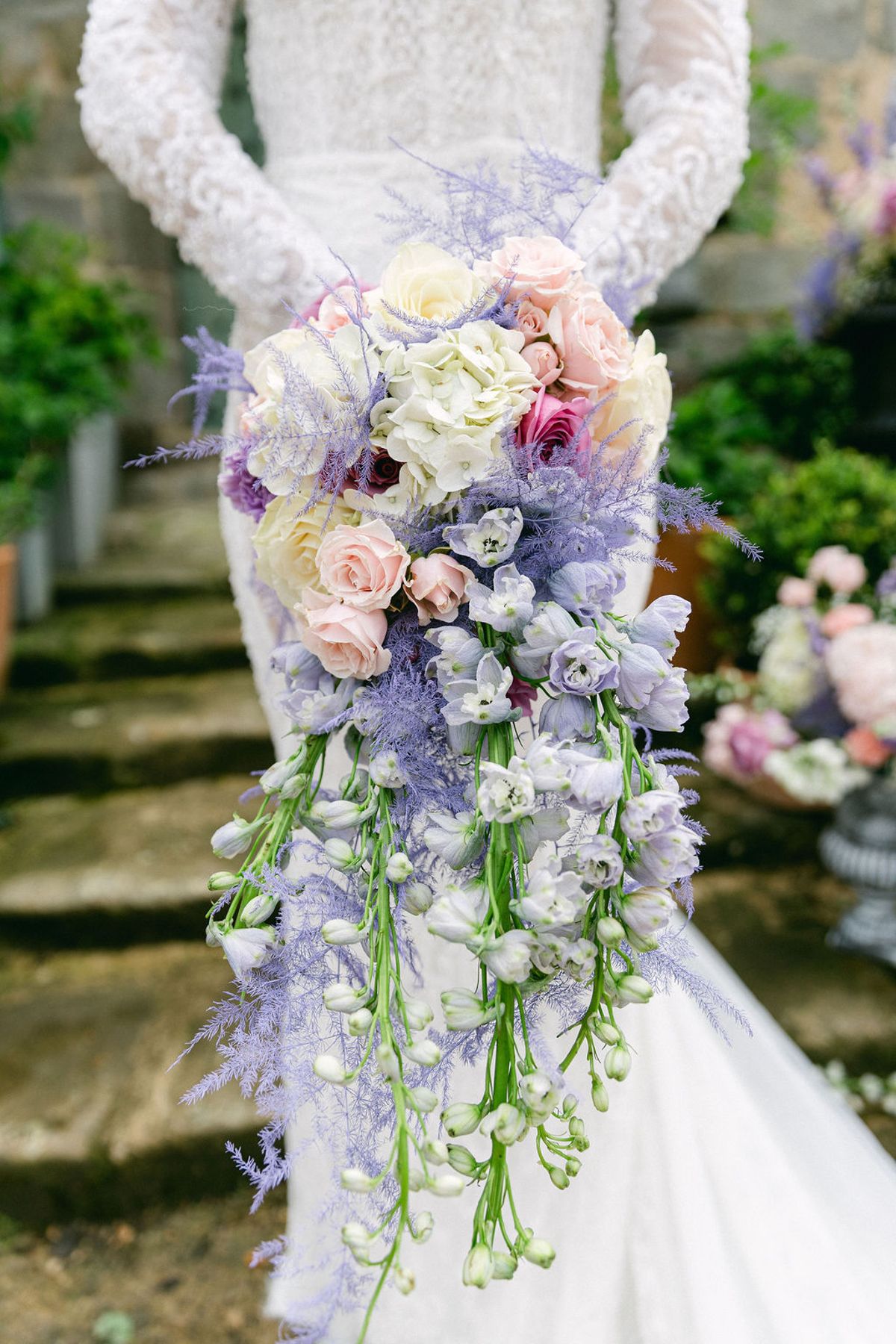 A cascading bouquet of cream, pink and lilac flowers held behind the back of a bride in a lace gown.