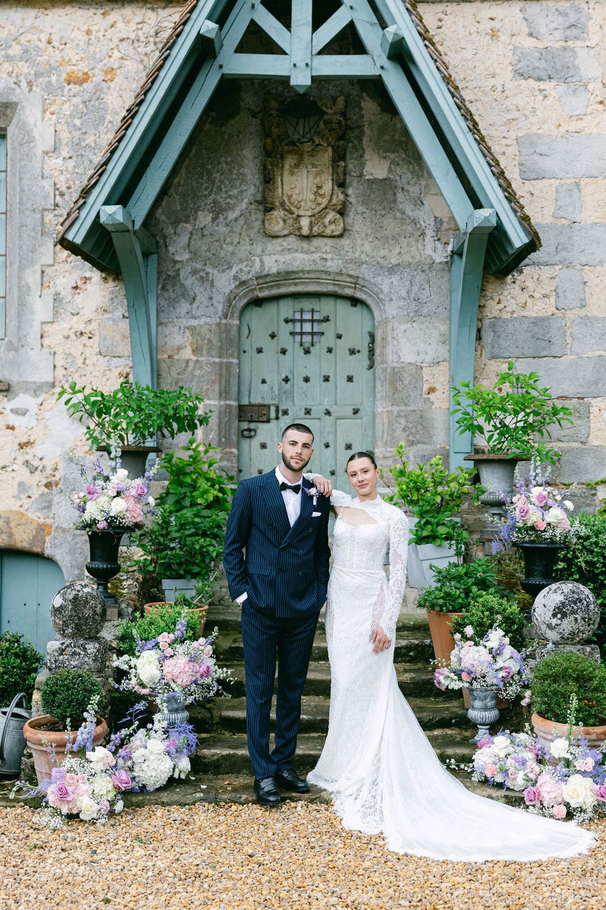 The groom in a navy suit and the bride in a long-sleeved lace gown stand on steps surrounded by pastel flowers and potted plants.