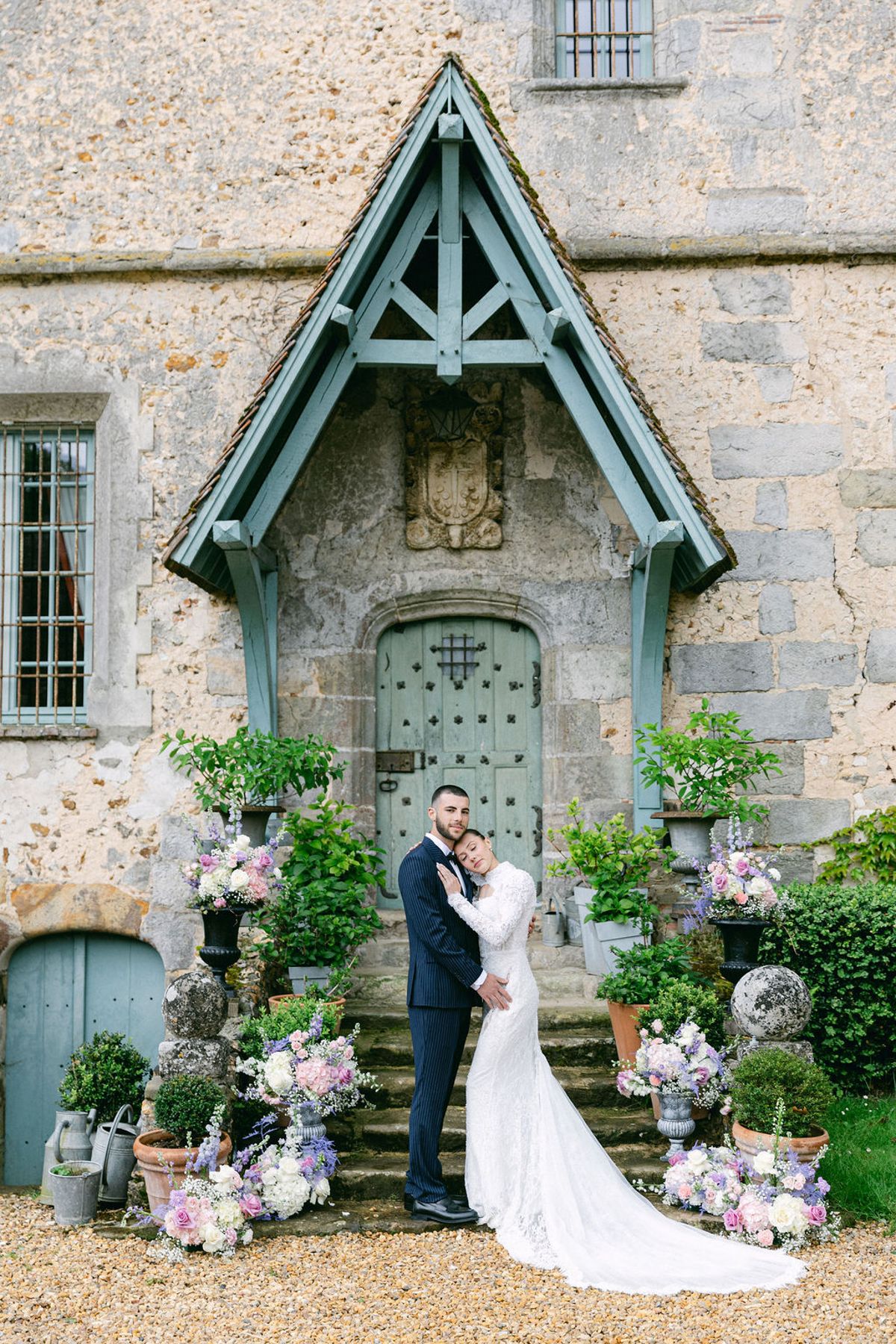 The couple embrace on stone steps in front of a chateau door, surrounded by pots of pink and lilac flowers.