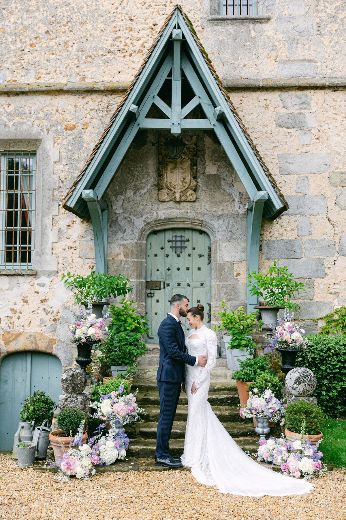 The couple stand face to face on stone steps in front of a pale blue door, surrounded by flowers and potted plants.