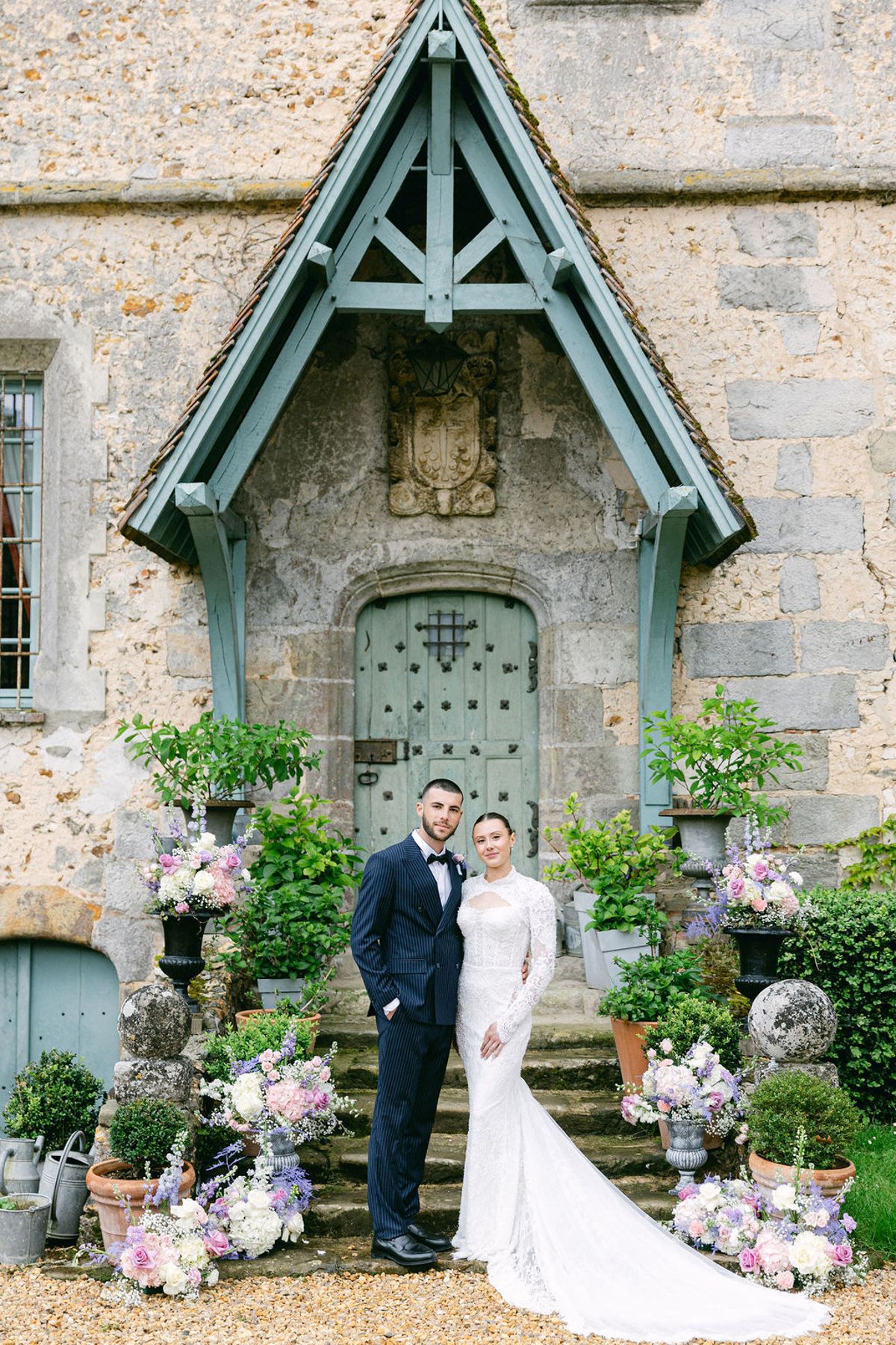 The groom in a navy suit and the bride in a long-sleeved lace gown stand before a stone chateau door surrounded by flowers.