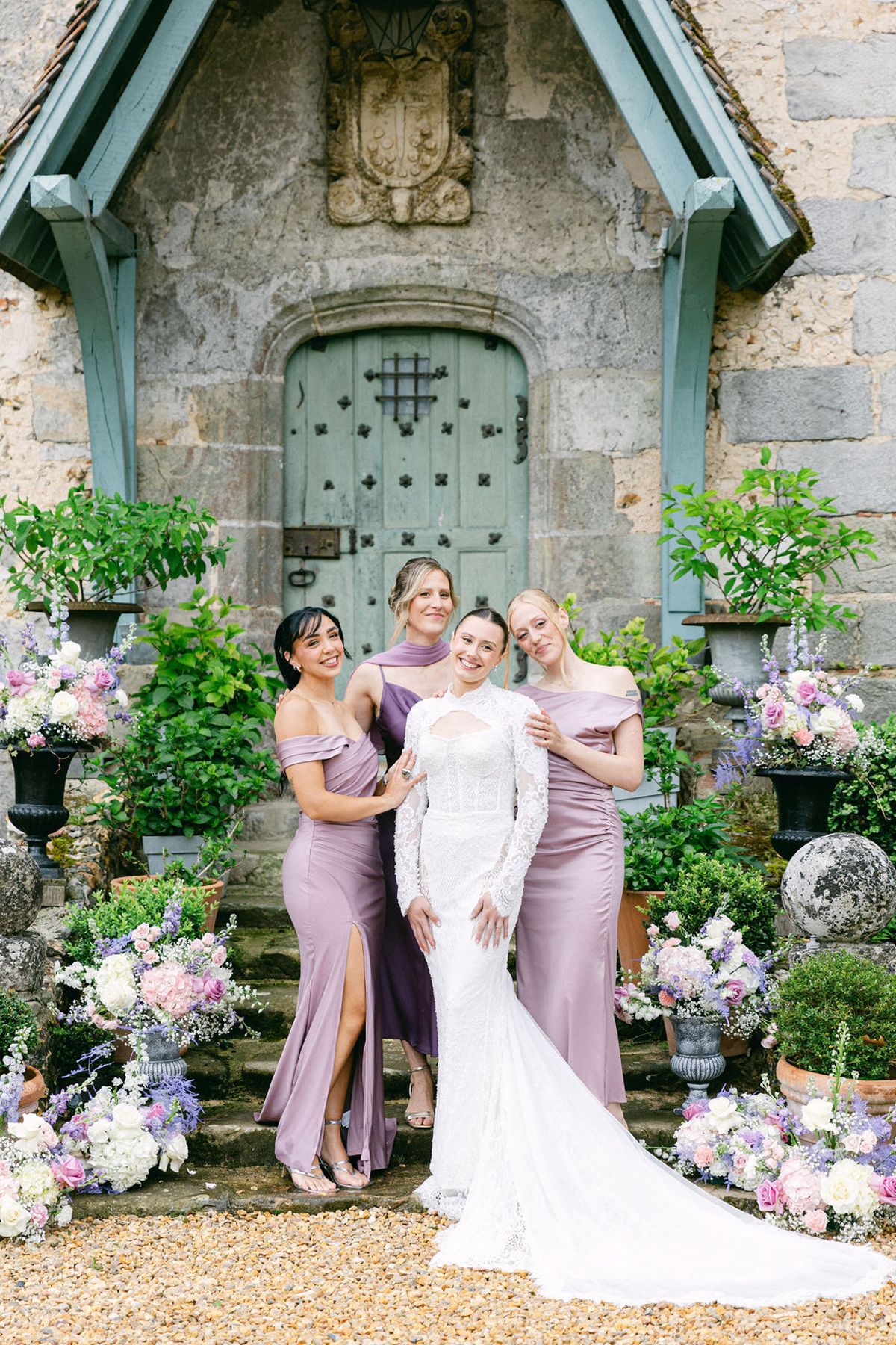 The bride and three bridesmaids in mauve dresses pose close together on stone steps in front of a green door, surrounded by flowers.