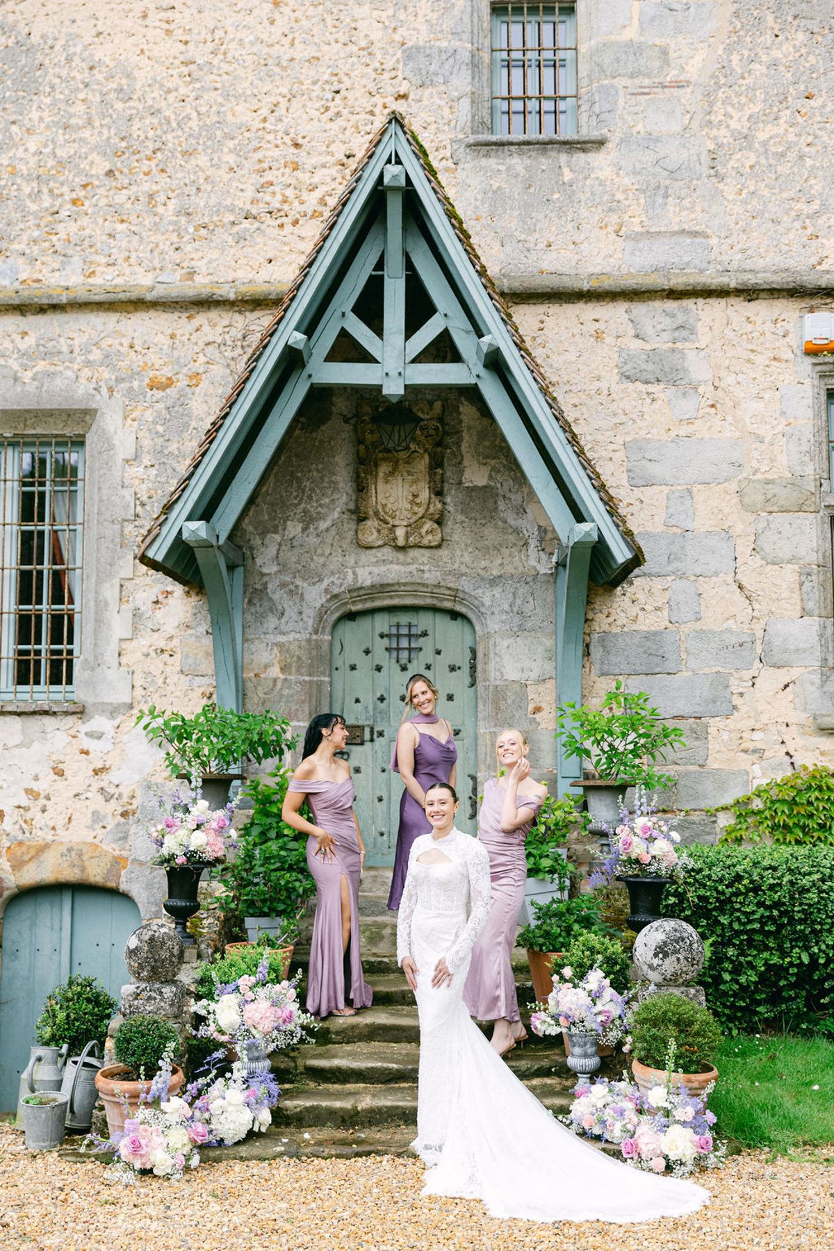 The bride in a lace gown stands on stone steps with three bridesmaids in mauve dresses beneath a pale blue porch, surrounded by flowers.