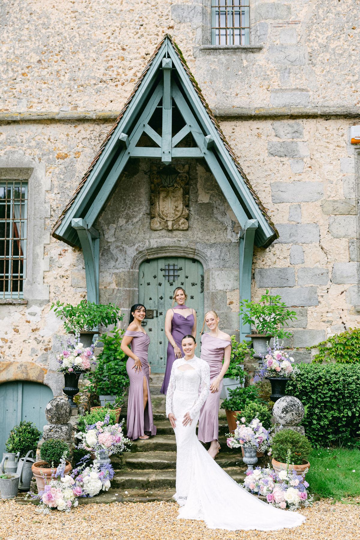 The bride stands on stone steps in front of a chateau door with three bridesmaids in lilac satin dresses behind her.