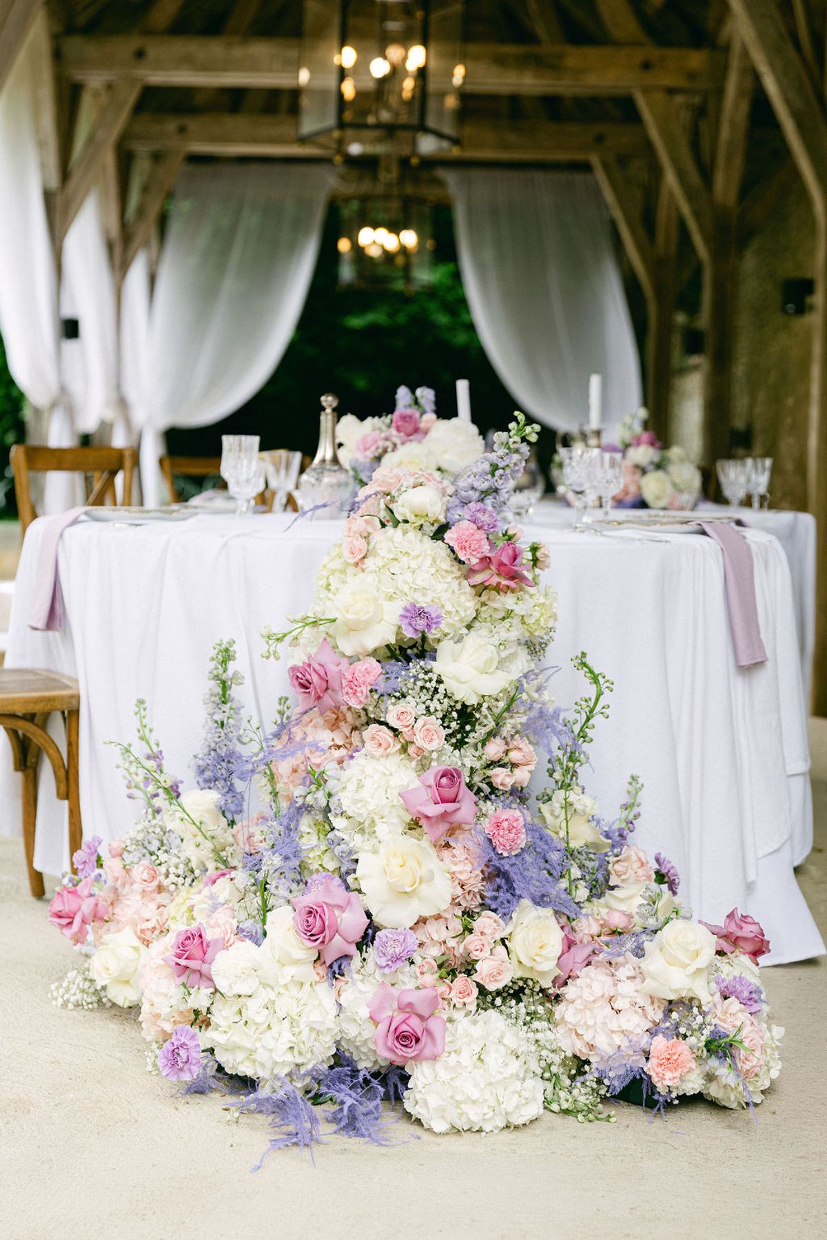 A cascade of pink, white and lilac flowers spills across the floor in front of a white-clothed table set with candles, inside a timber barn.