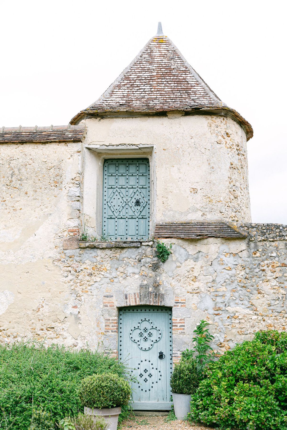 A weathered stone building with a round tower, a pale blue window and door, and clipped box hedges in front.