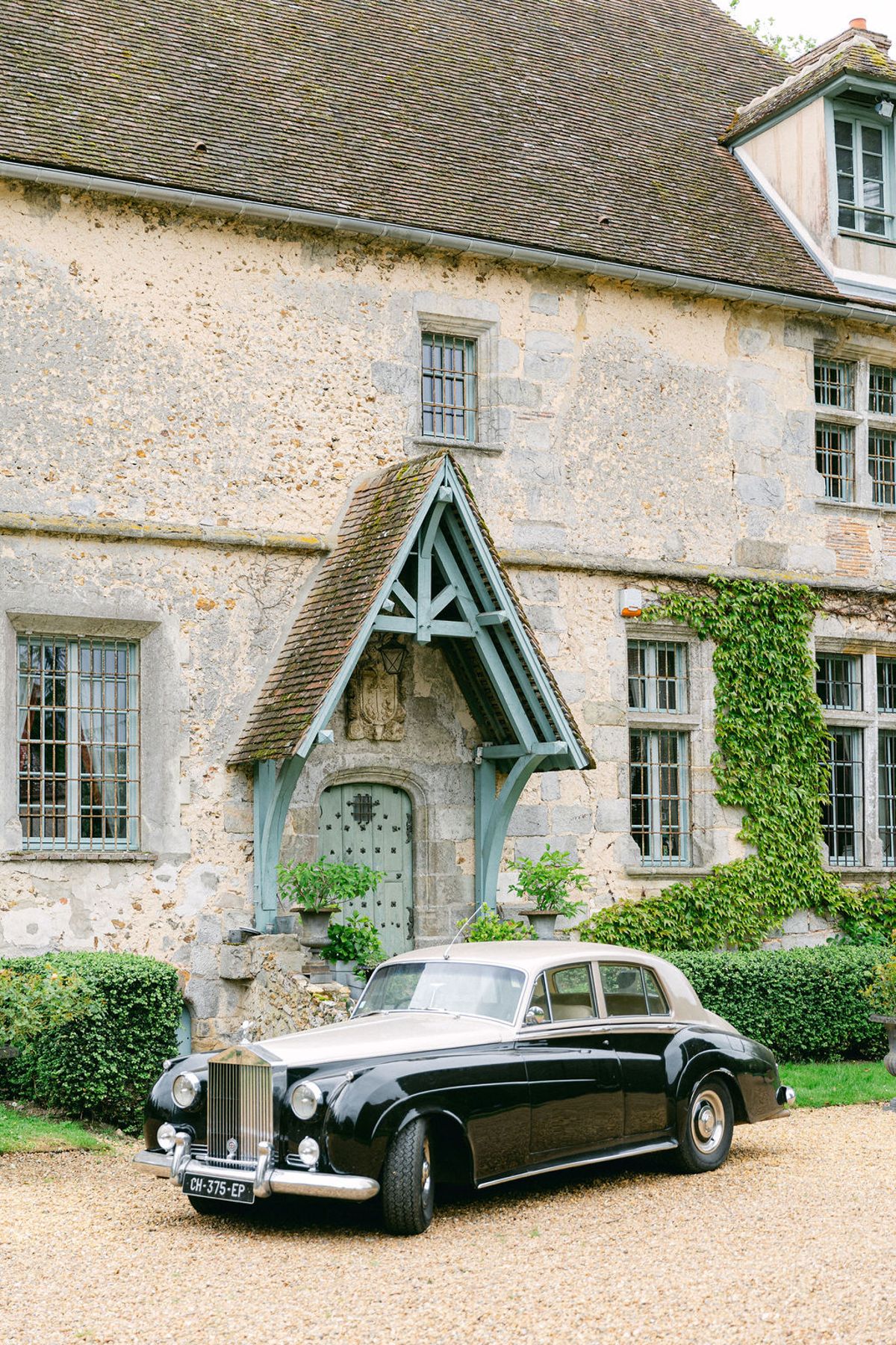 A vintage black car is parked on gravel in front of a stone chateau with ivy, a timber porch and tall windows.