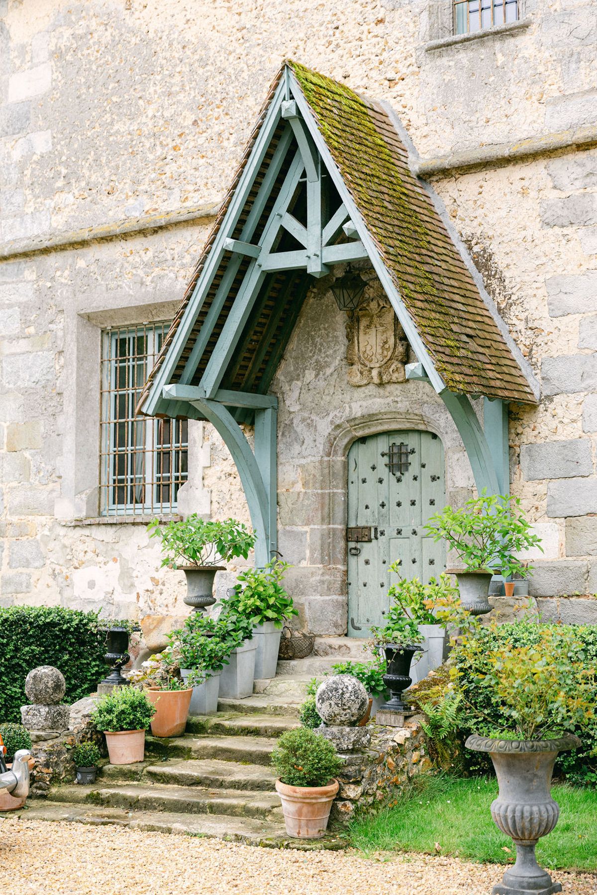 A stone building with a pointed, moss-covered porch roof, a green door and stone steps lined with potted plants and urns.