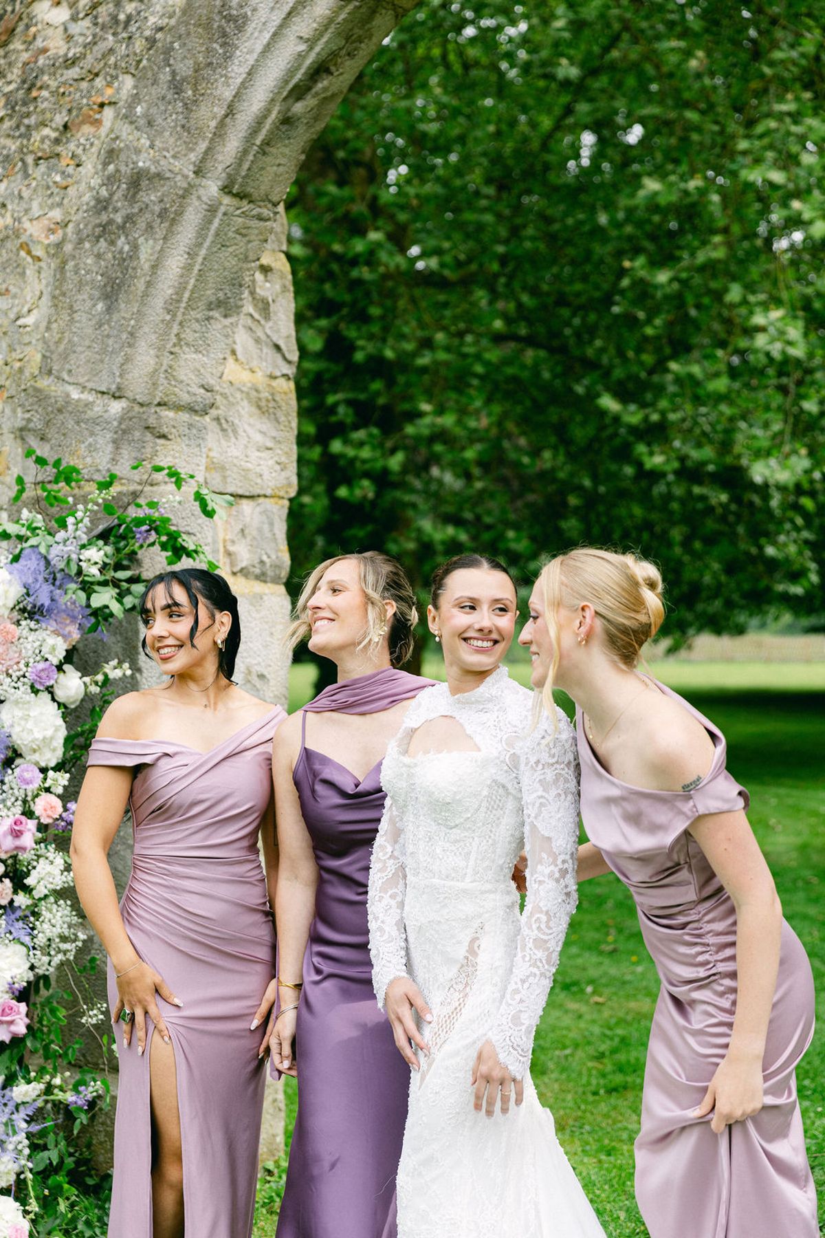 The bride and three bridesmaids in lilac satin dresses stand together on a lawn, laughing and leaning towards one another beside a stone arch.