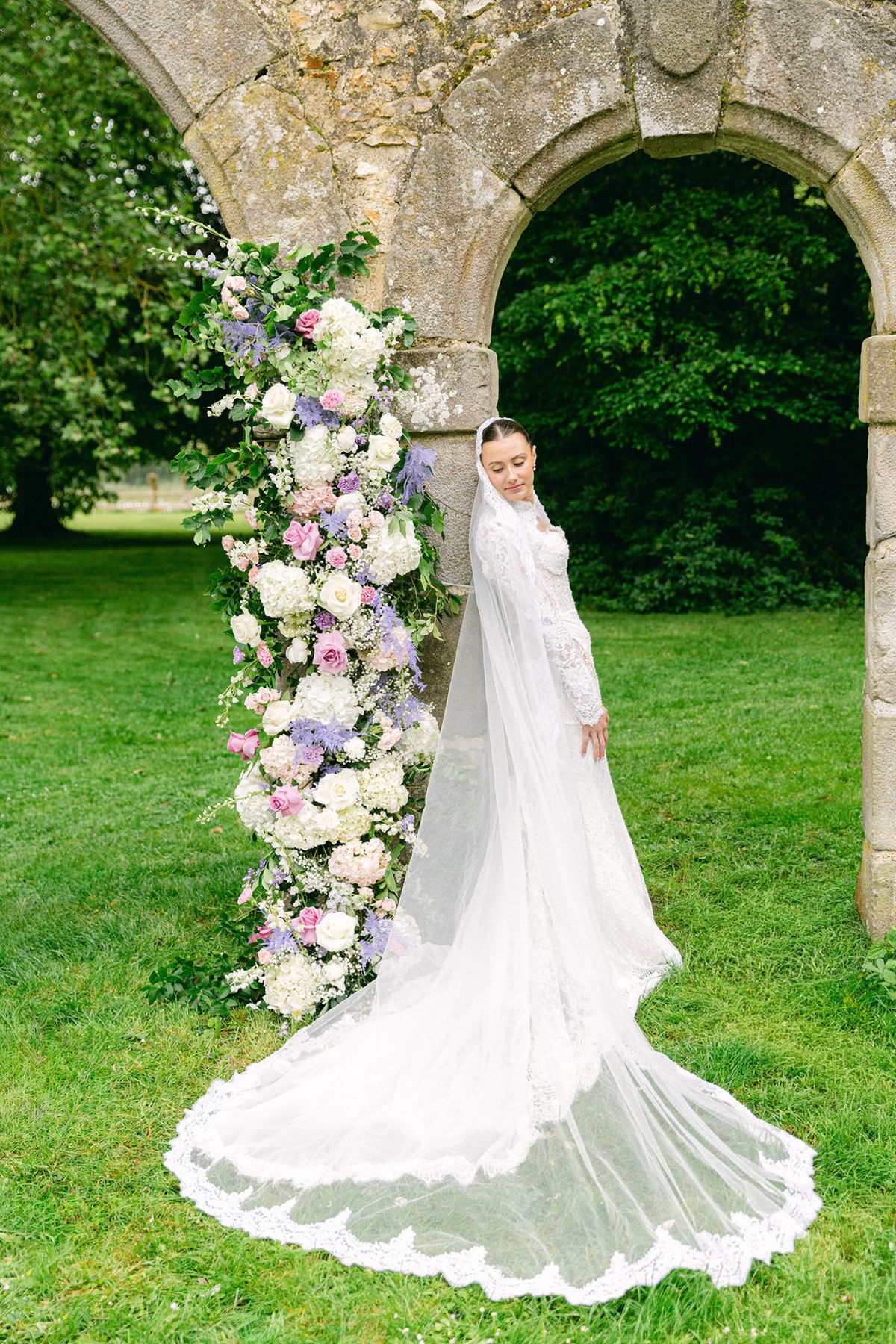 The bride stands in profile on a lawn beside a tall pastel floral arrangement, her long lace-edged veil trailing behind her before a stone arch.