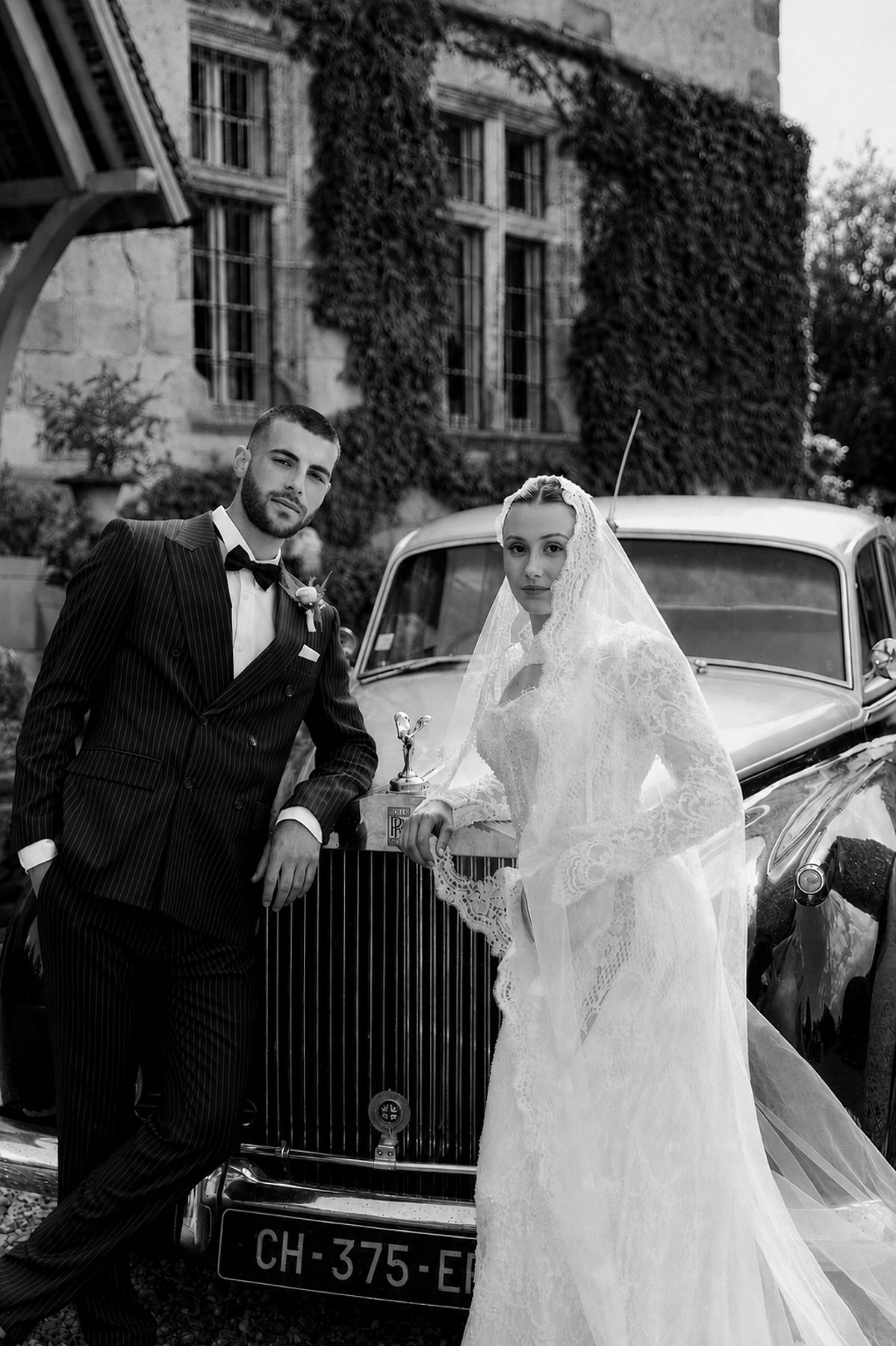 In black and white, the groom in a dark suit and the bride in a veil lean on the front of a vintage car.