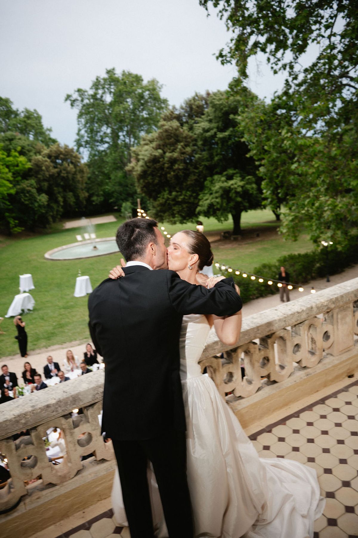 Clara and Constantin embrace on the balustrade terrace beside the fountain as evening lights come on