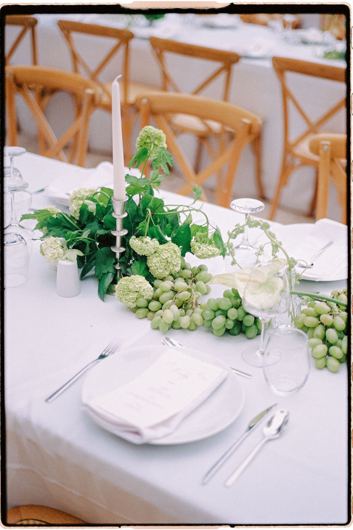 A reception table set with candles, greenery and grapes trailing across white linen