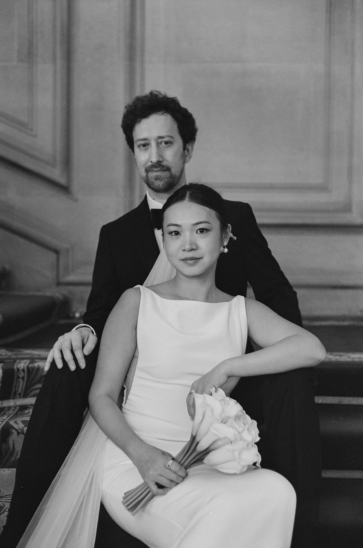 In black and white, the bride sits holding a bouquet while the groom sits behind her with his hand on her arm.