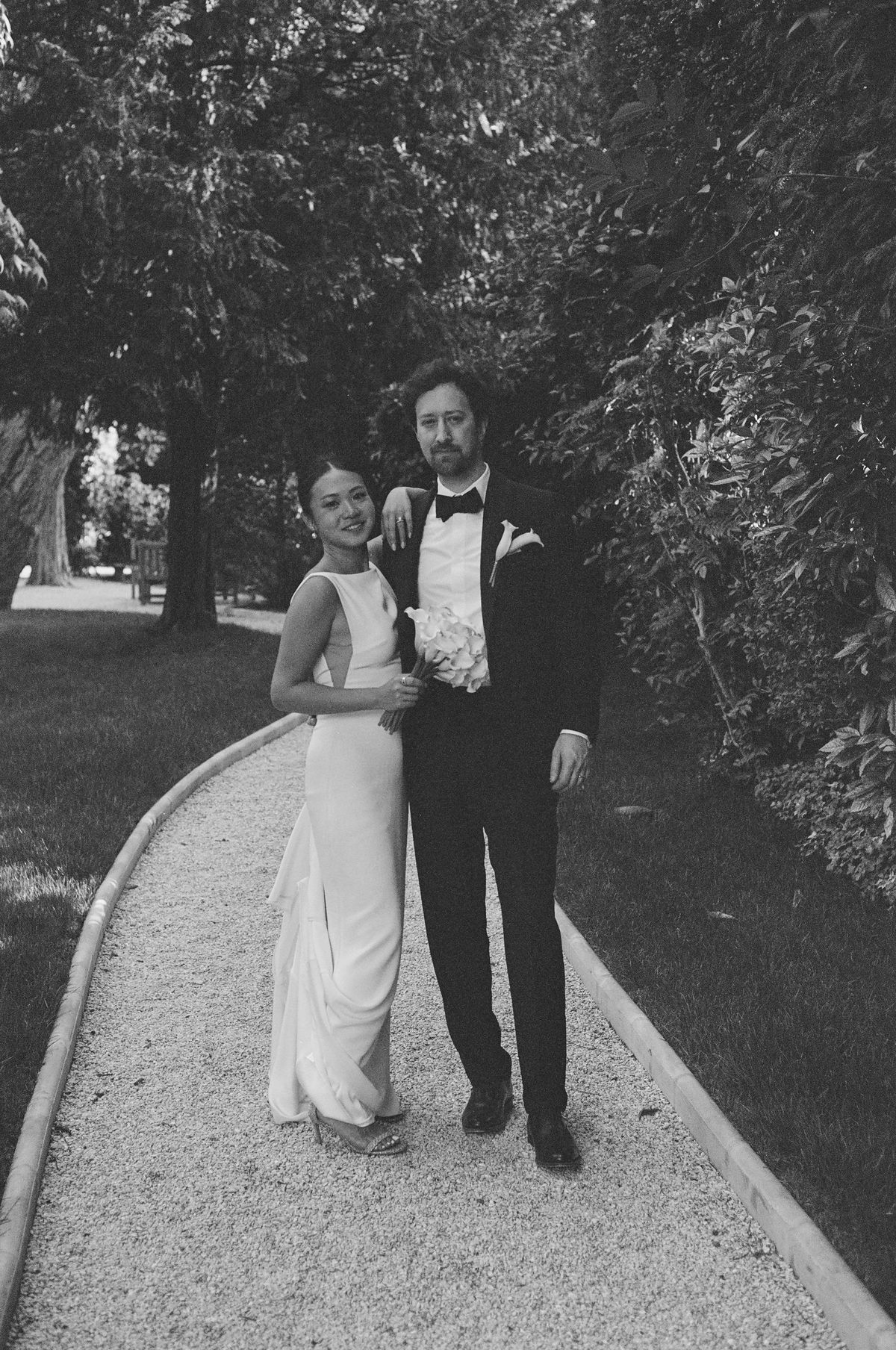 In black and white, the couple walk side by side along a pale gravel path lined with trees, the groom in a tuxedo.