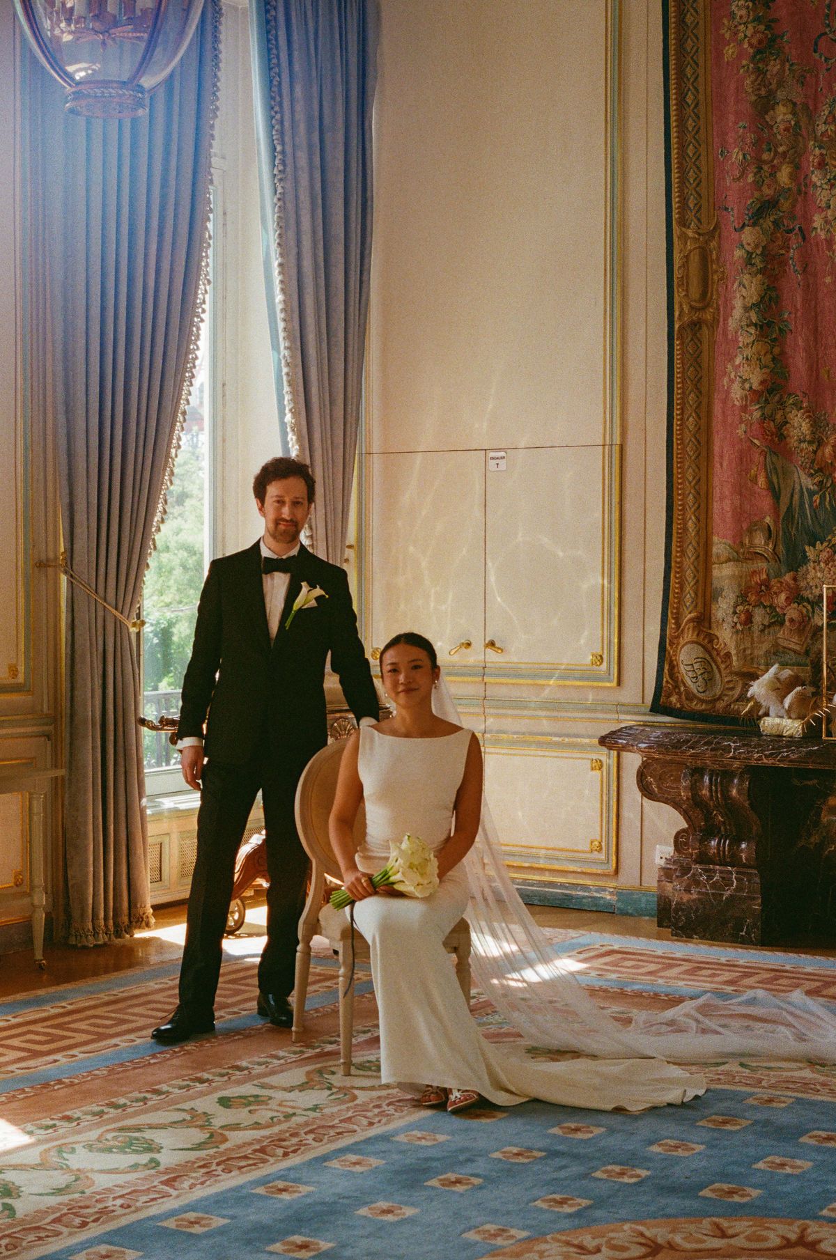 The groom in a dark suit stands beside the bride, who sits in a white dress with a veil, near tall blue curtains.